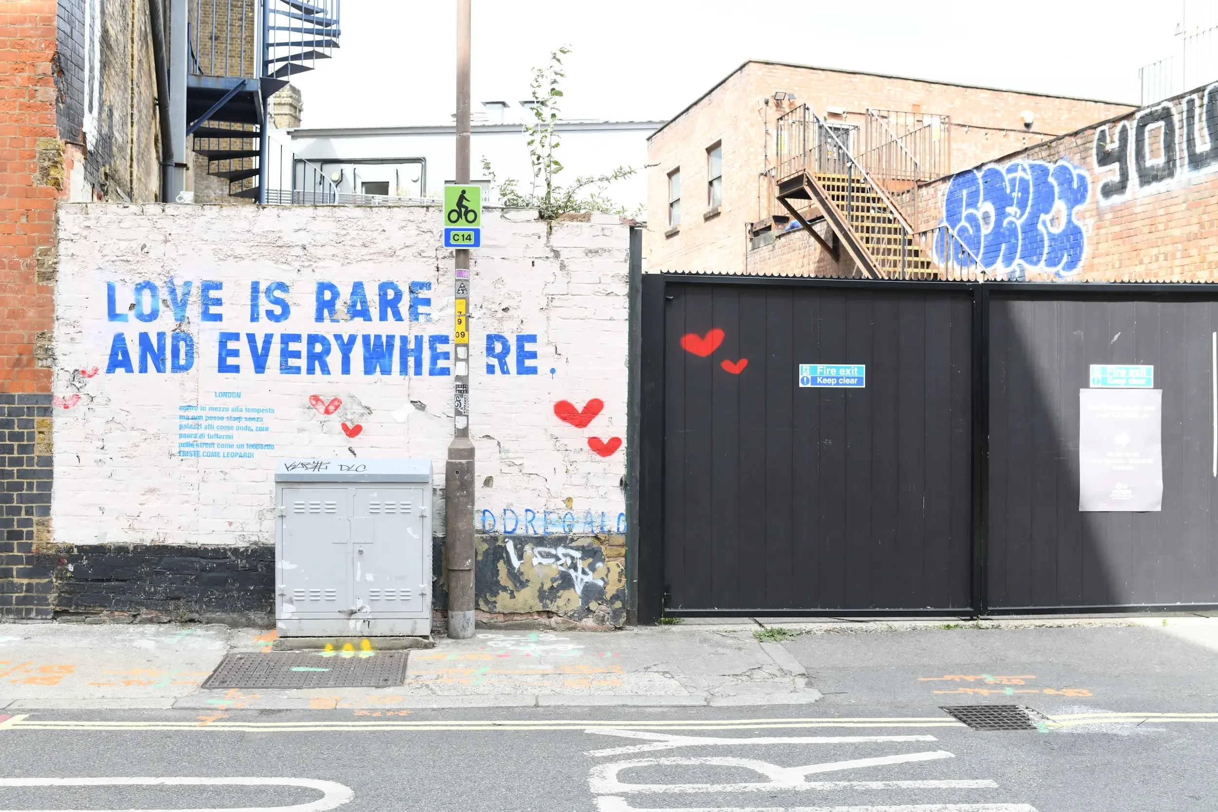 Urban alleyway with graffiti, a white wall with blue text that reads 'Love is rare and everywhere,' a black gate, a fire exit sign, and a metal staircase on a brick building.