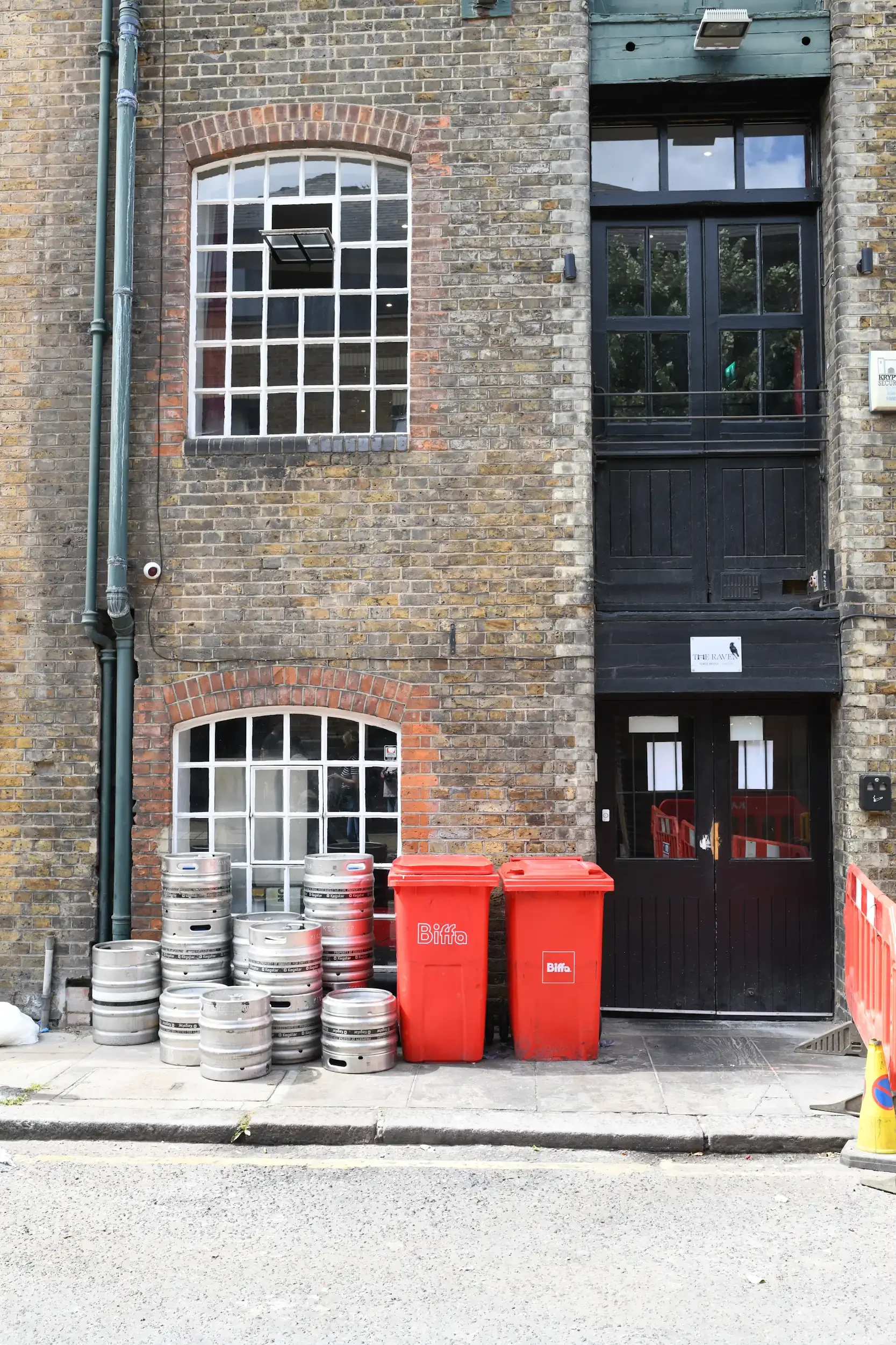 A brick building with three large windows and a black door. Several beer kegs and two red trash bins are placed in front of the building. A street with a sidewalk is visible in the foreground.