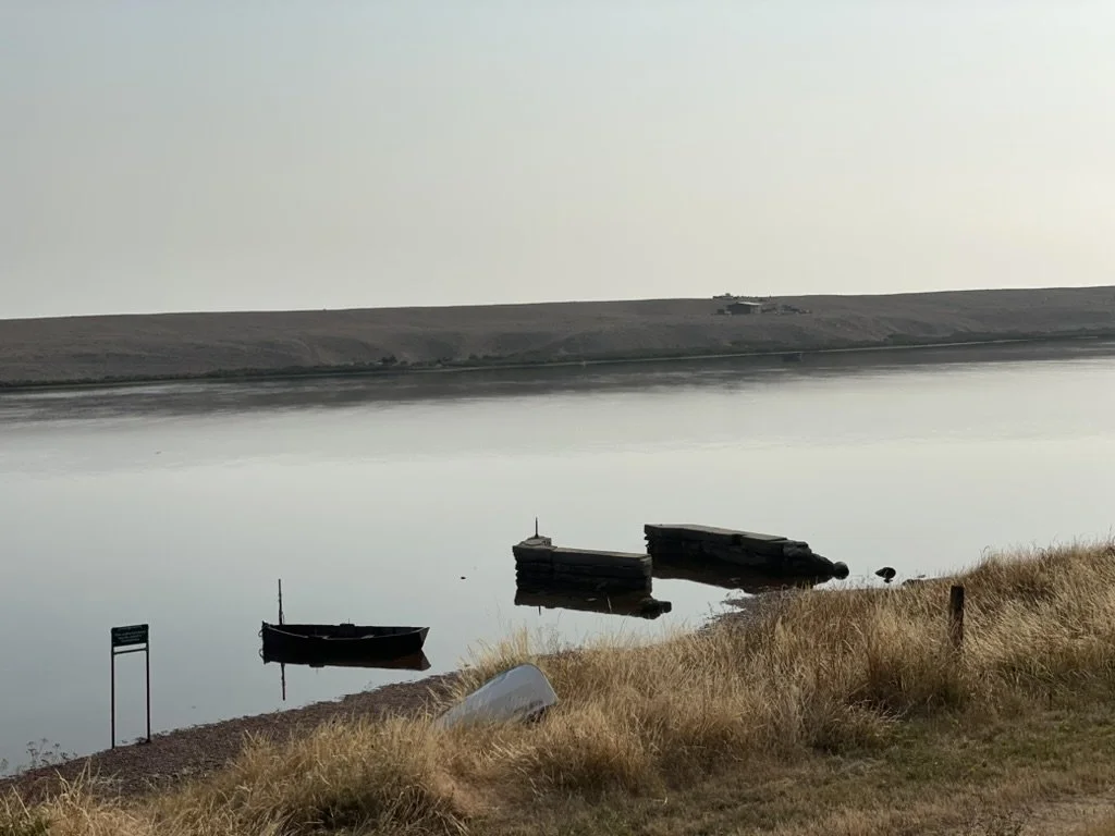 Calm water body with a small boat floating near the shore, some concrete structures in the water, dry grass on the shoreline, and a distant hill or landmass in the background.