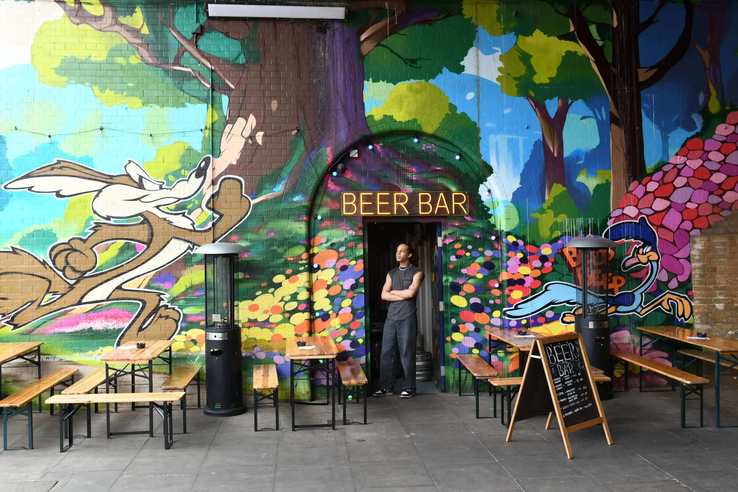 Indoor scene of a beer bar with colorful mural artwork on brick wall, featuring trees and cartoon characters, neon sign that says 'BEER BAR', and a person standing in front of the artwork. There are wooden tables and benches set up in front of the mu