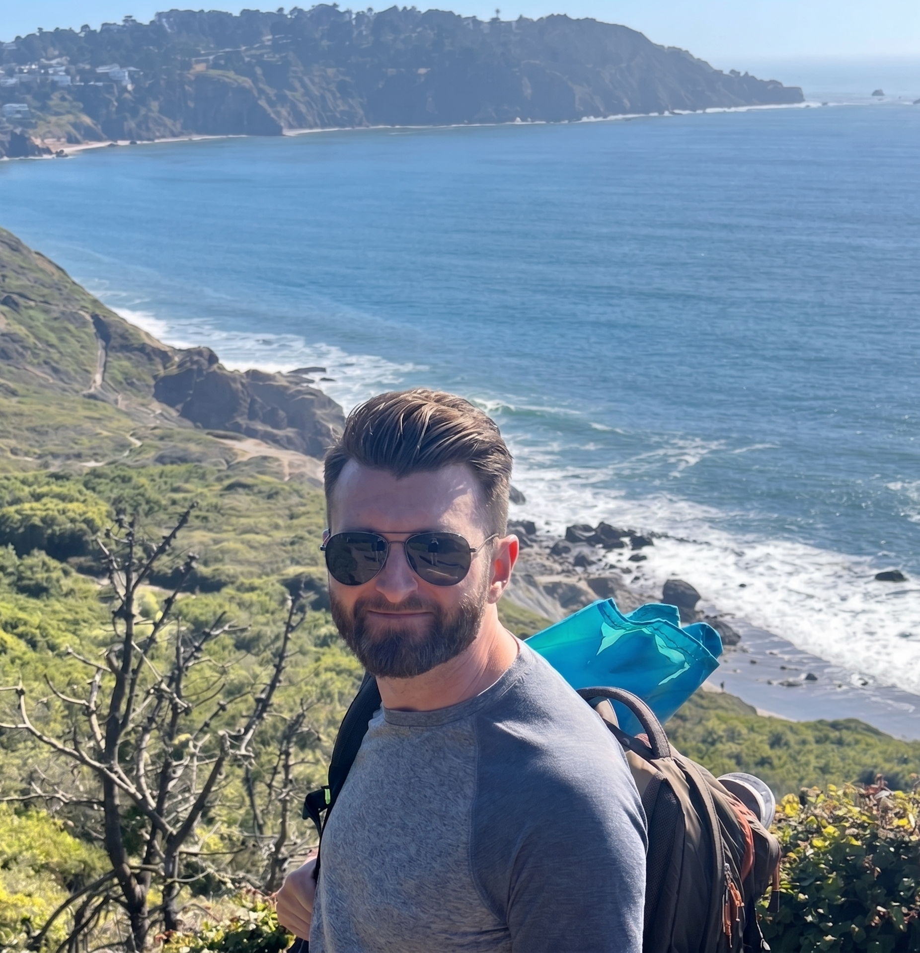 A man with sunglasses and a beard taking a selfie outdoors near the coast with cliffs, ocean, and houses in the background.