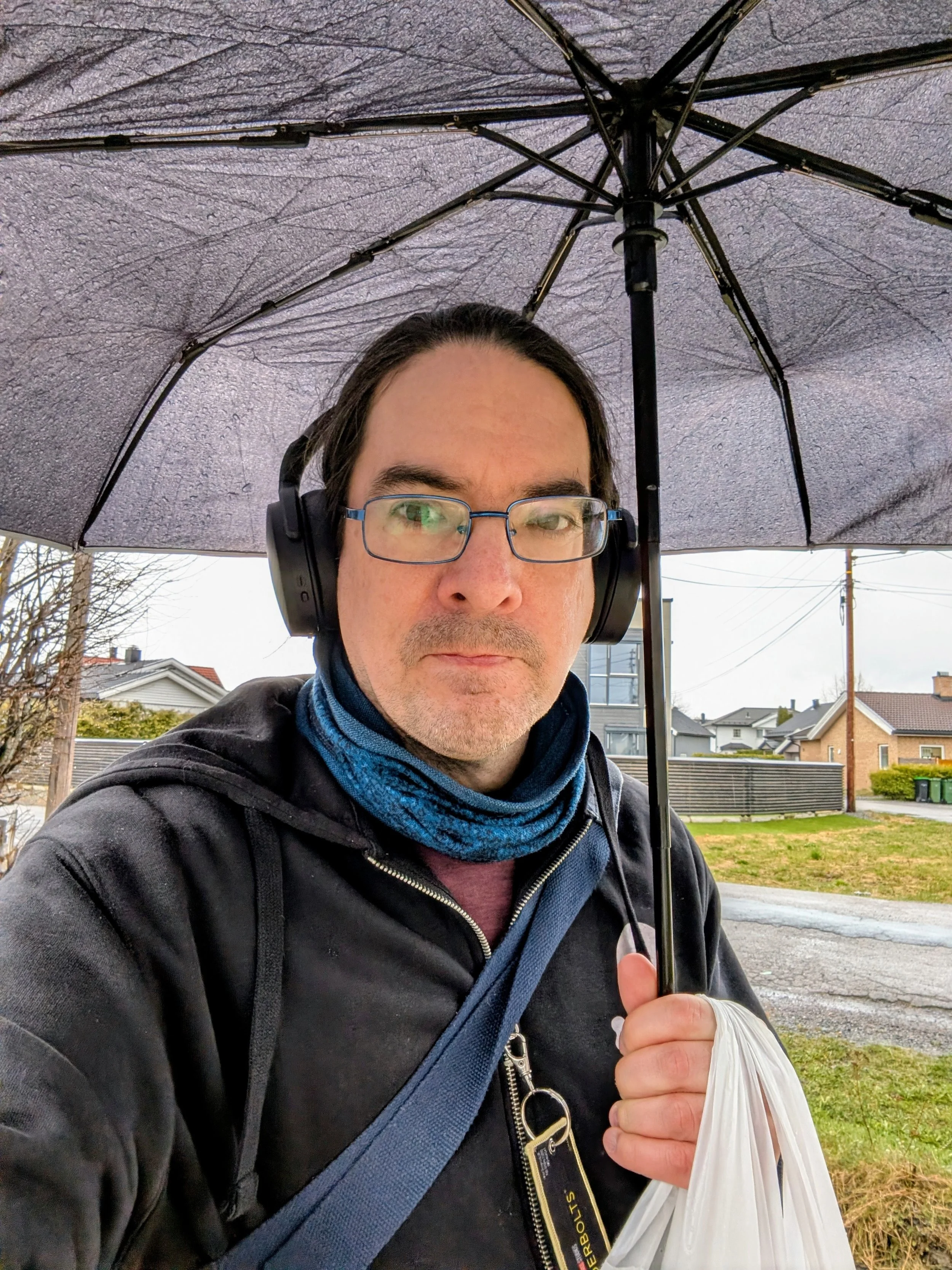 A man wearing glasses, headphones, and a black jacket is holding a black umbrella and a white plastic bag on a city street with houses and power lines in the background.