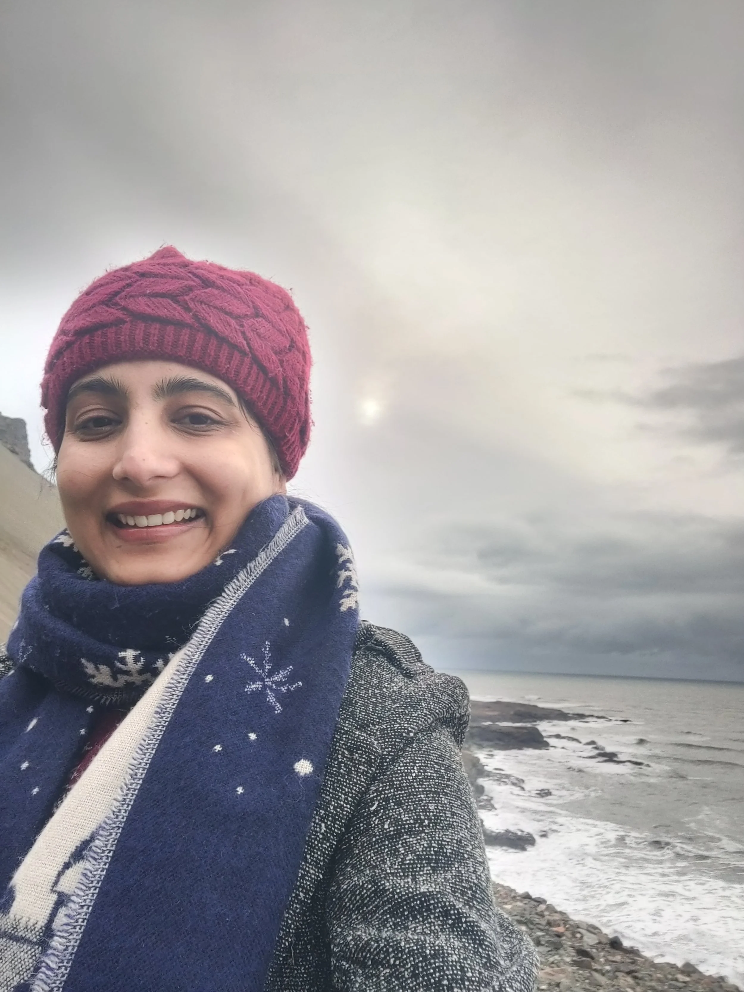 Person smiling outdoors on a rocky shoreline with cloudy sky and ocean in background, wearing a red knit hat and patterned scarf.