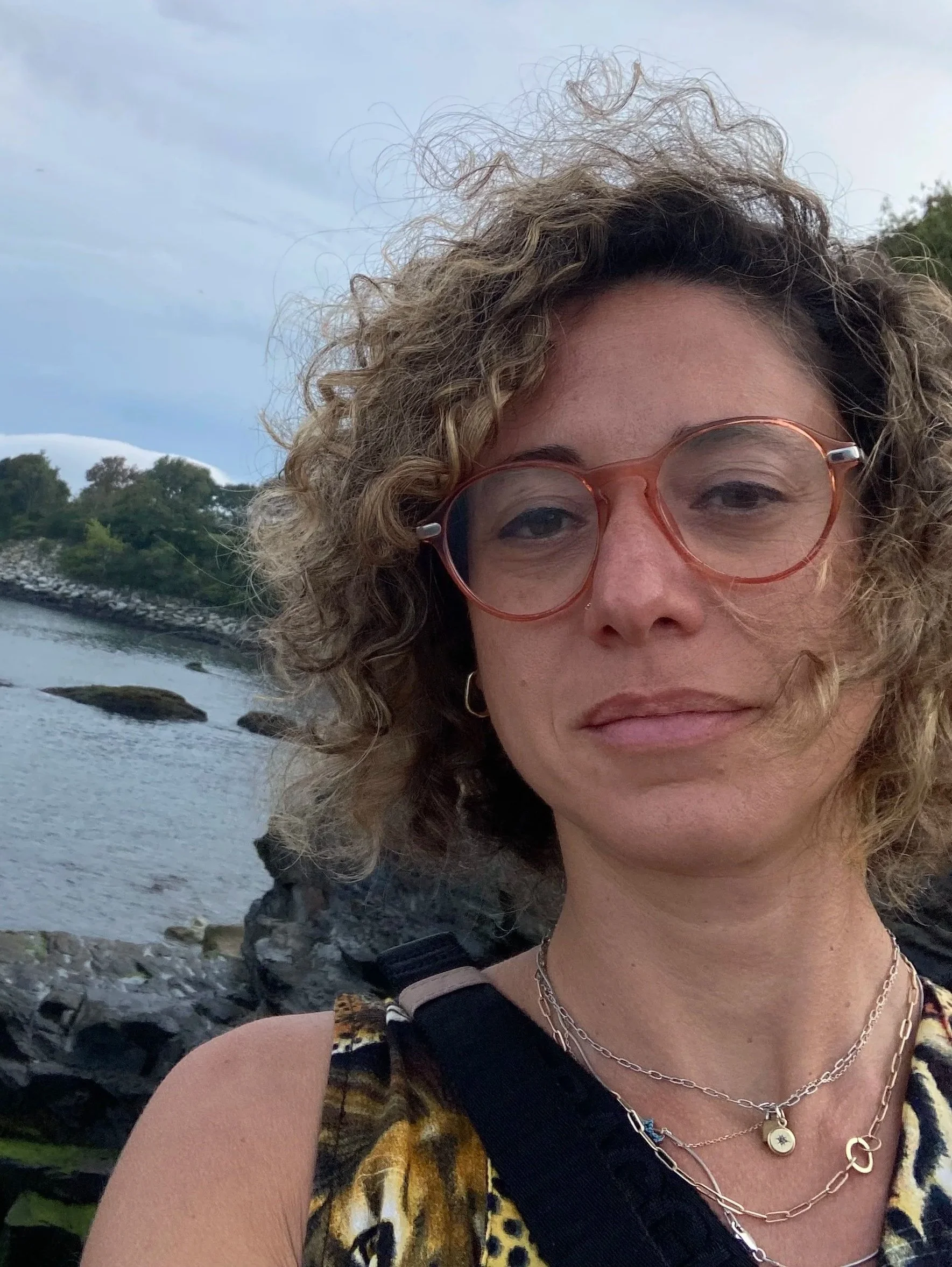 A woman with curly light brown hair and glasses taking a selfie outdoors near a body of water, with rocks and greenery in the background.