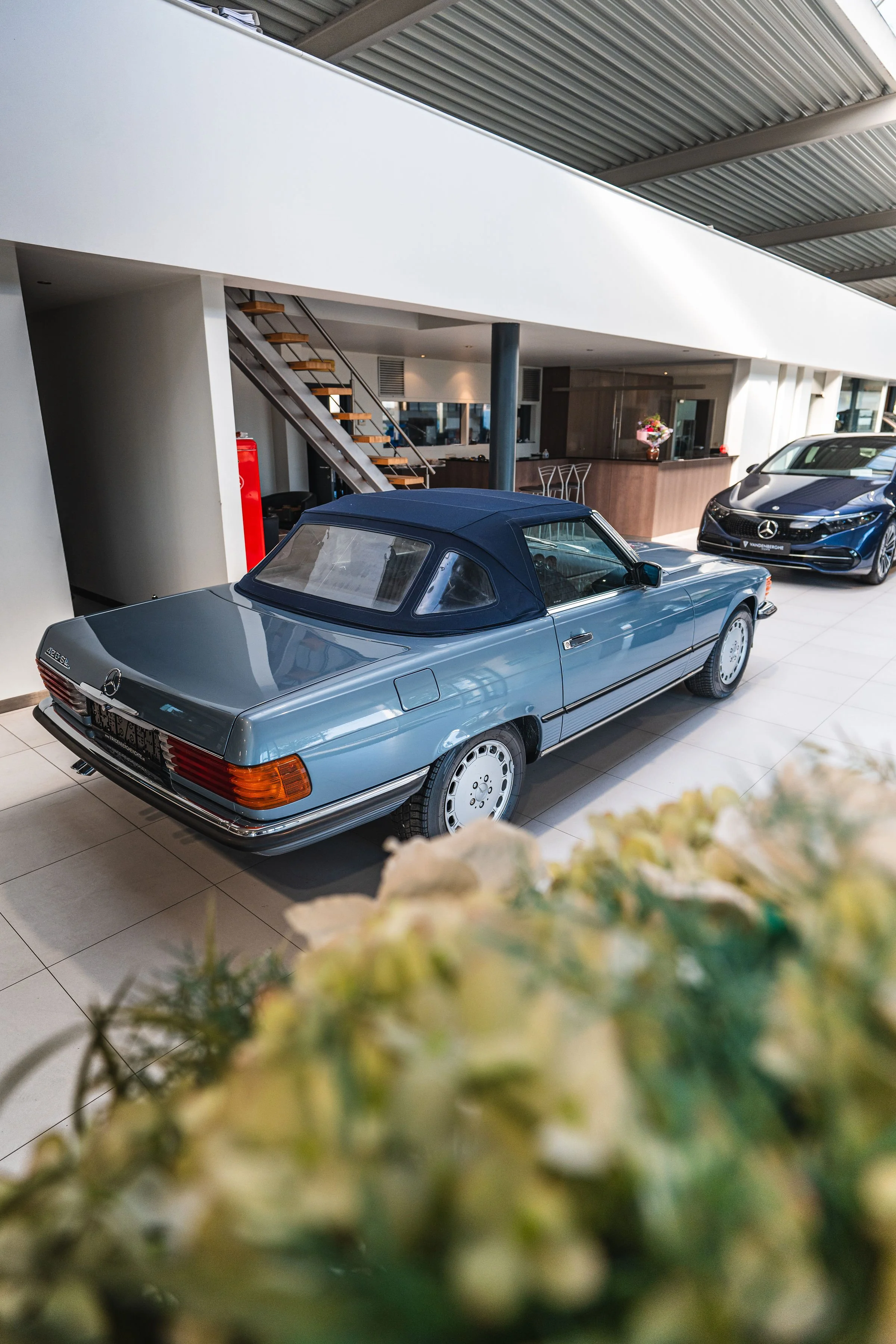 Witte klassieke Mercedes-Benz cabriolet in een autodealer of opslagruimte, onder een metalen plafond met spothouders, naast andere auto's.