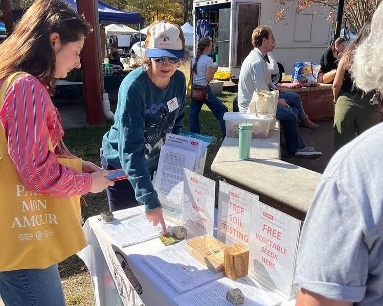 Photo of woman standing at table, talking to two people and pointing to soil and plant information sheets.