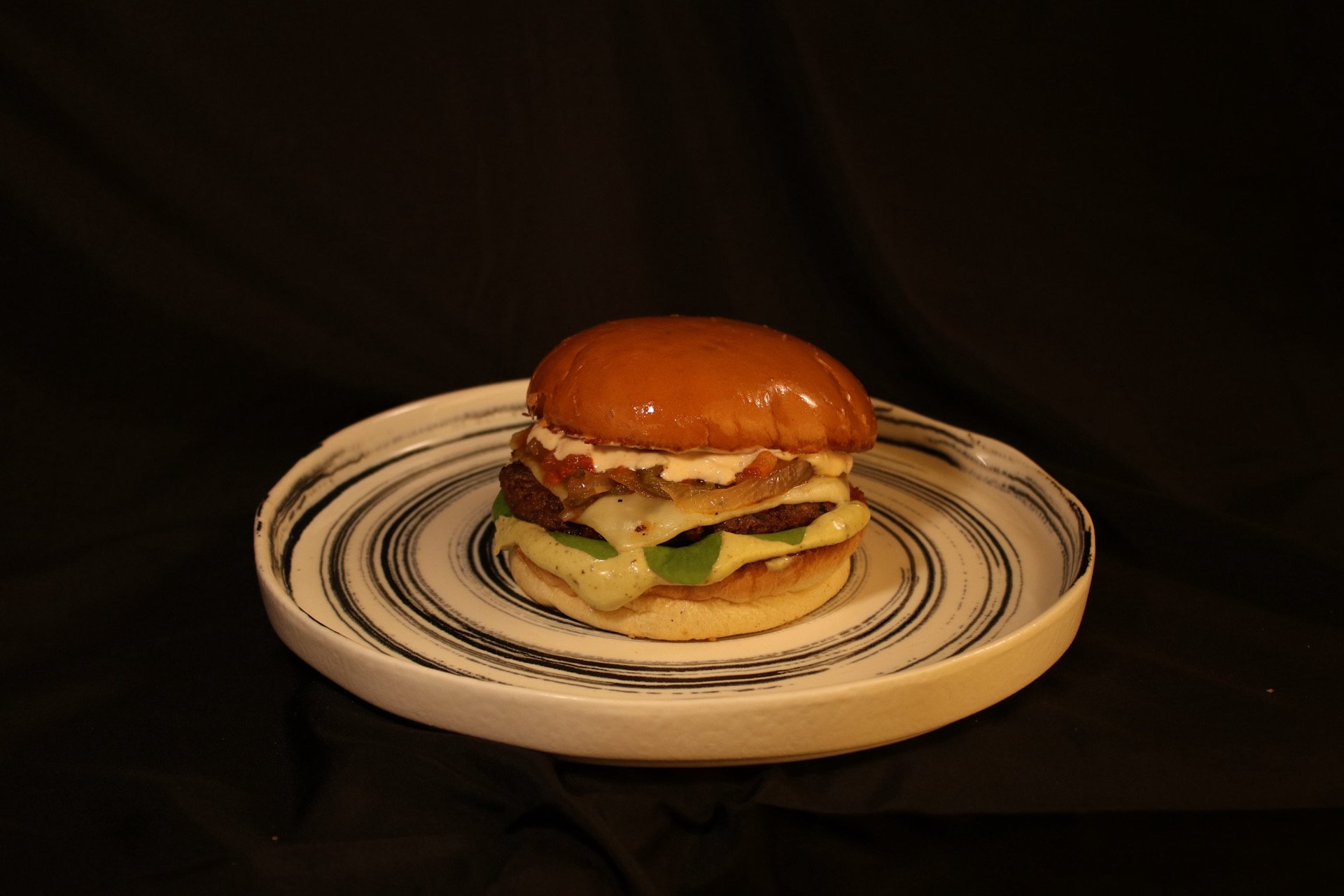 A gourmet burger with lettuce, cheese, bacon, a beef patty, and sauce on a decorative plate against a black background.