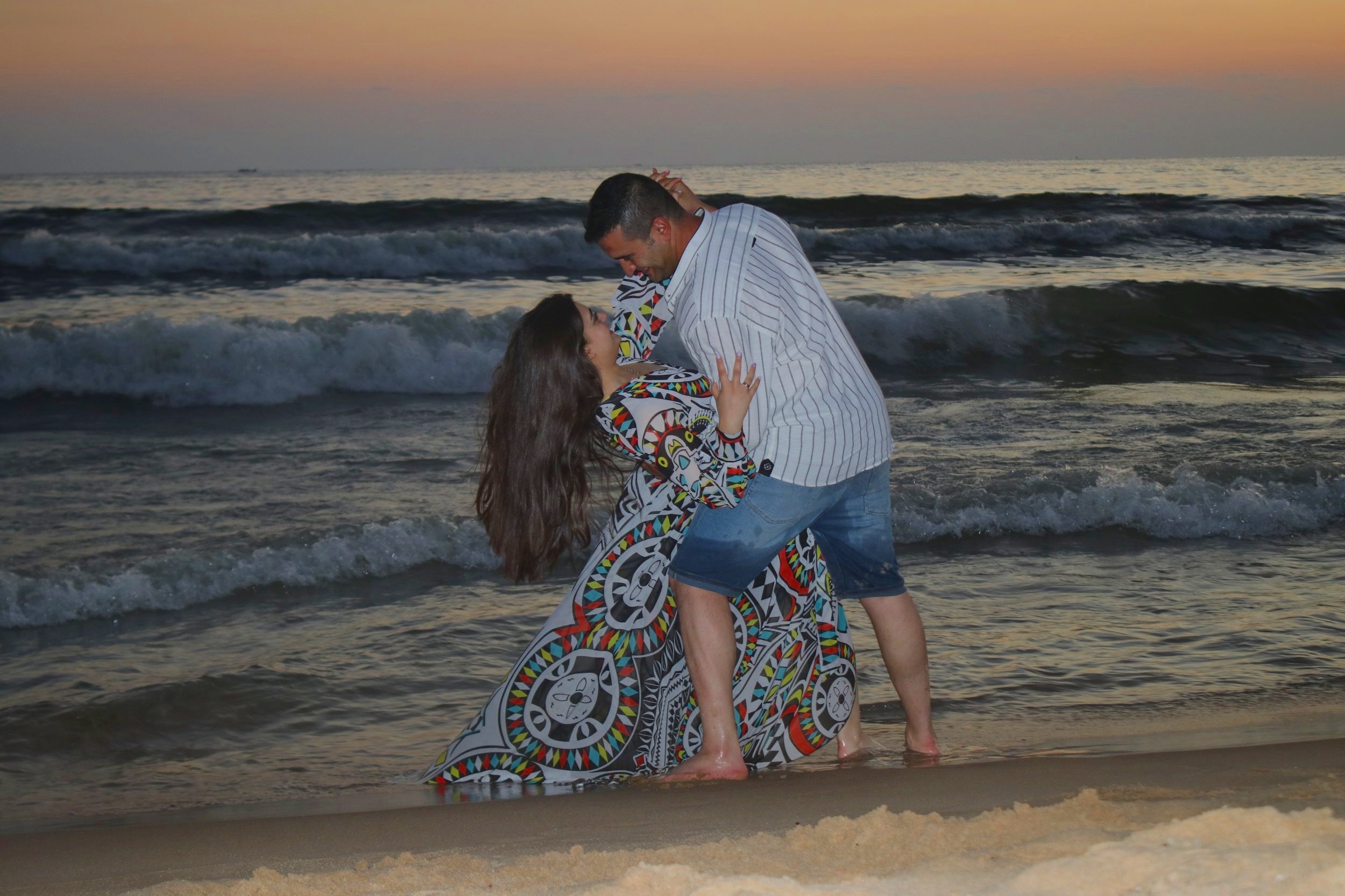 A couple is dancing on the beach at sunset, with waves in the background.