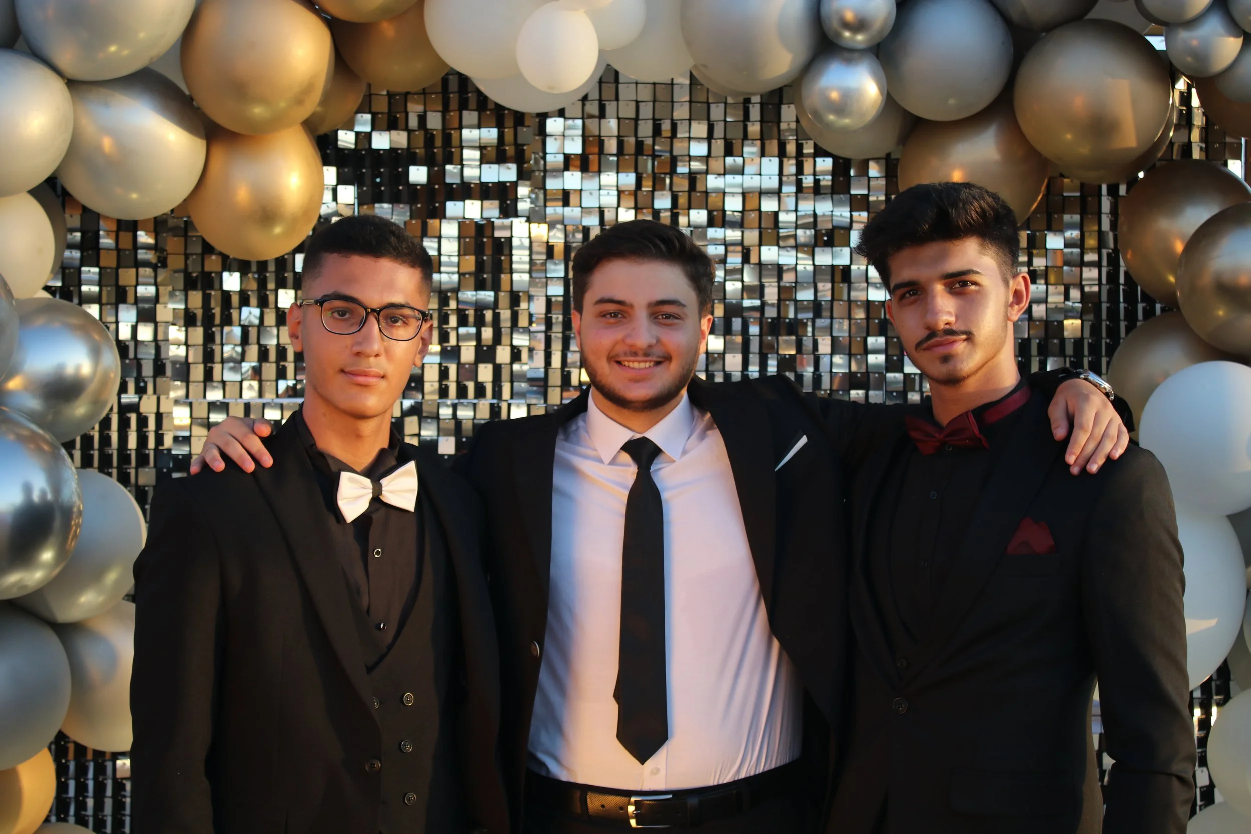 Three young men dressed in formal attire standing arm-in-arm at a celebratory event with balloon decorations behind them.