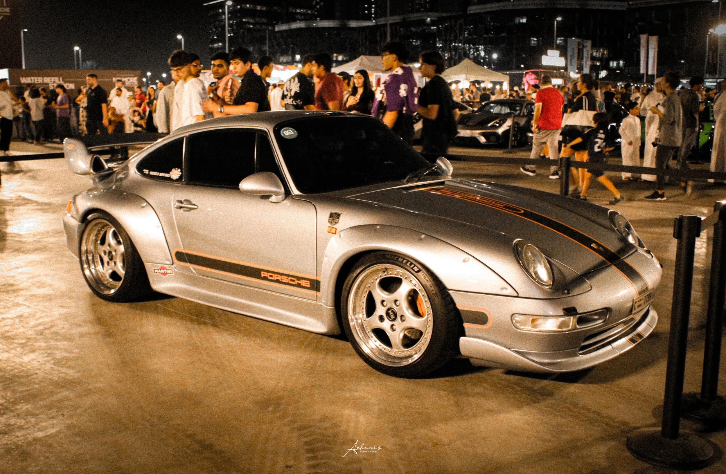 A silver Porsche 911 sports car with black and orange racing stripe details, parked at a nighttime car show with a crowd of people in the background.