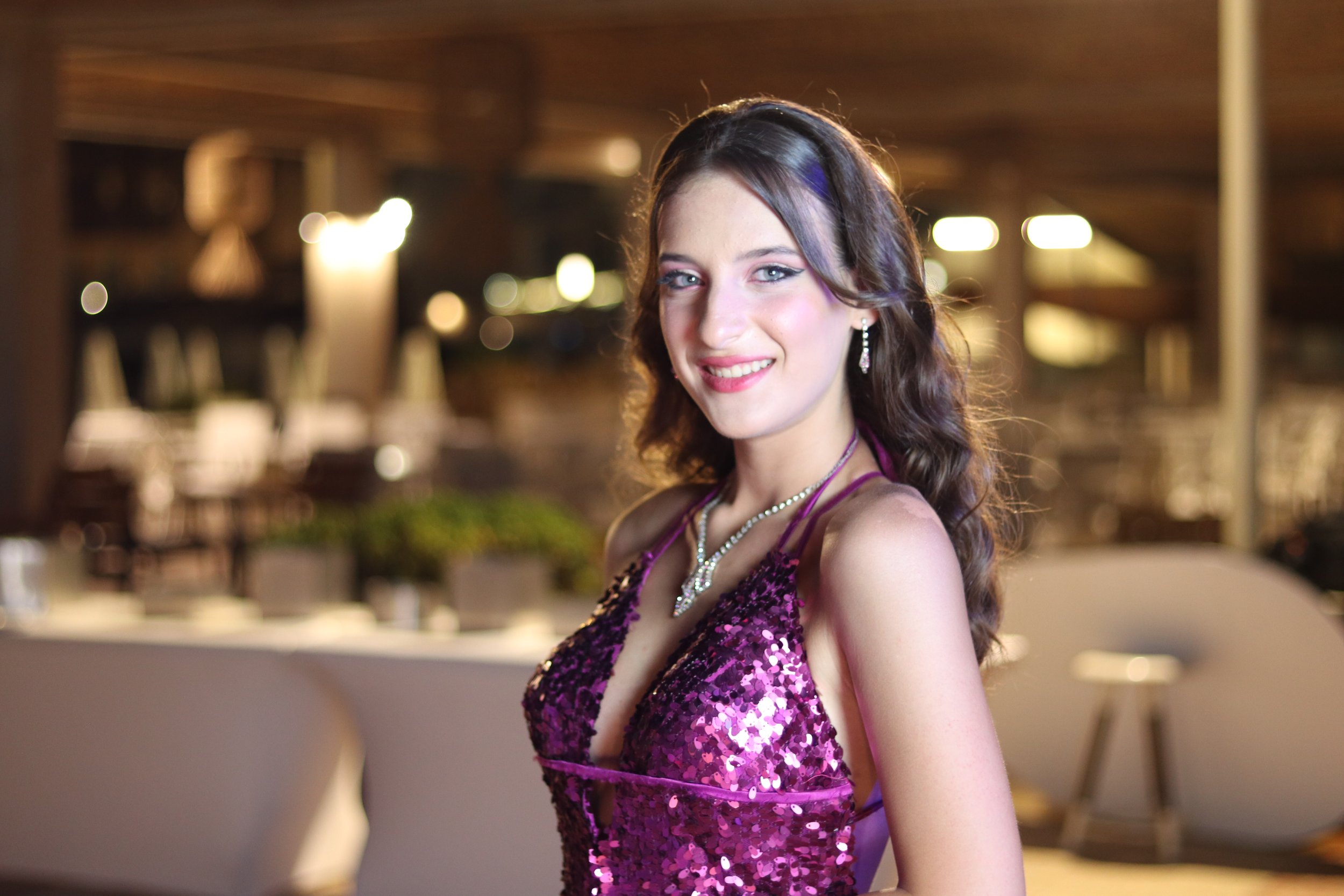 A young woman with long, wavy brown hair, wearing a purple, sequined dress and jewelry, smiling at the camera in a warm, indoor setting.