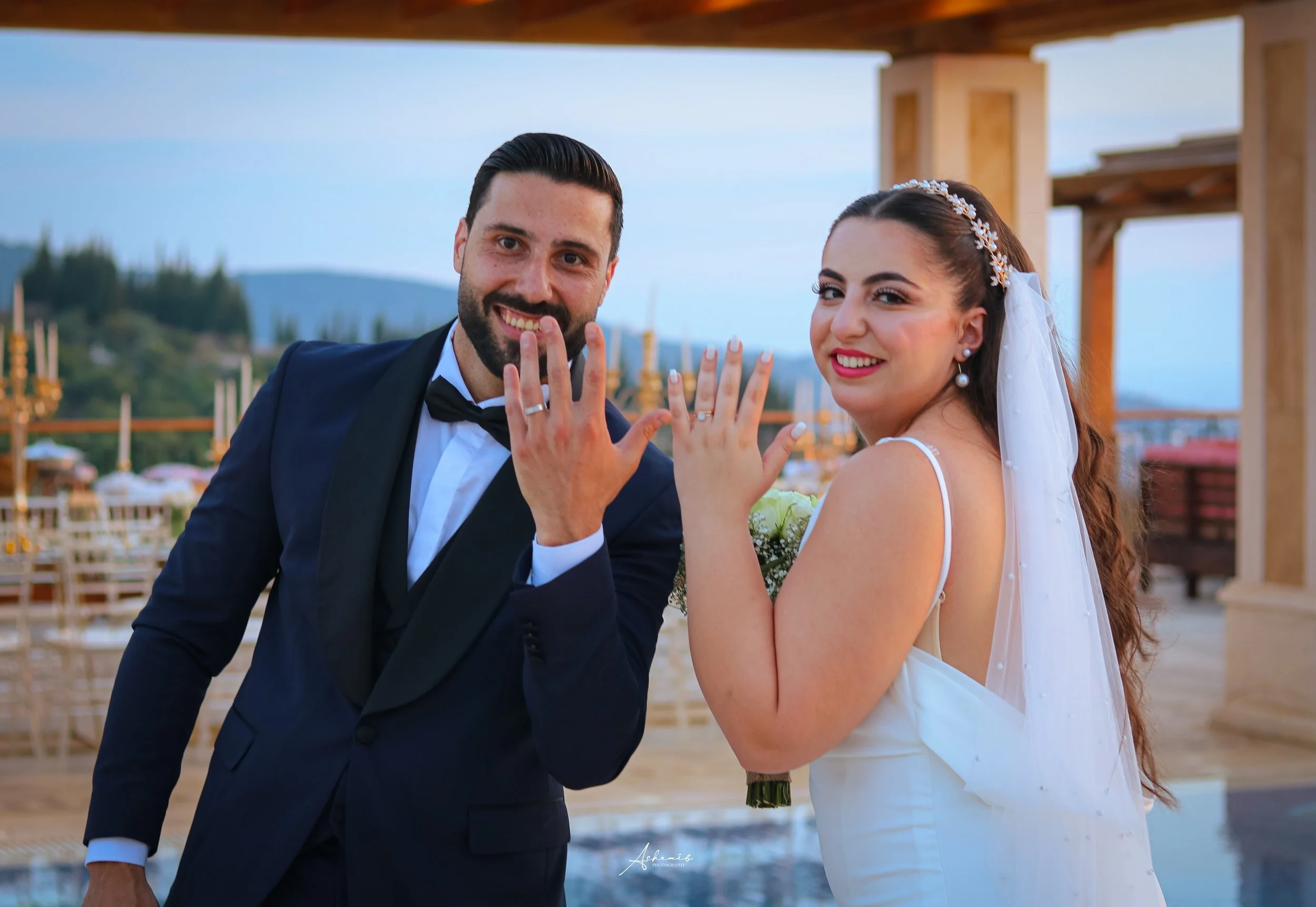 A newlywed couple smiling and showing their wedding rings, standing outdoors at sunset with a scenic backdrop, celebrating their marriage.