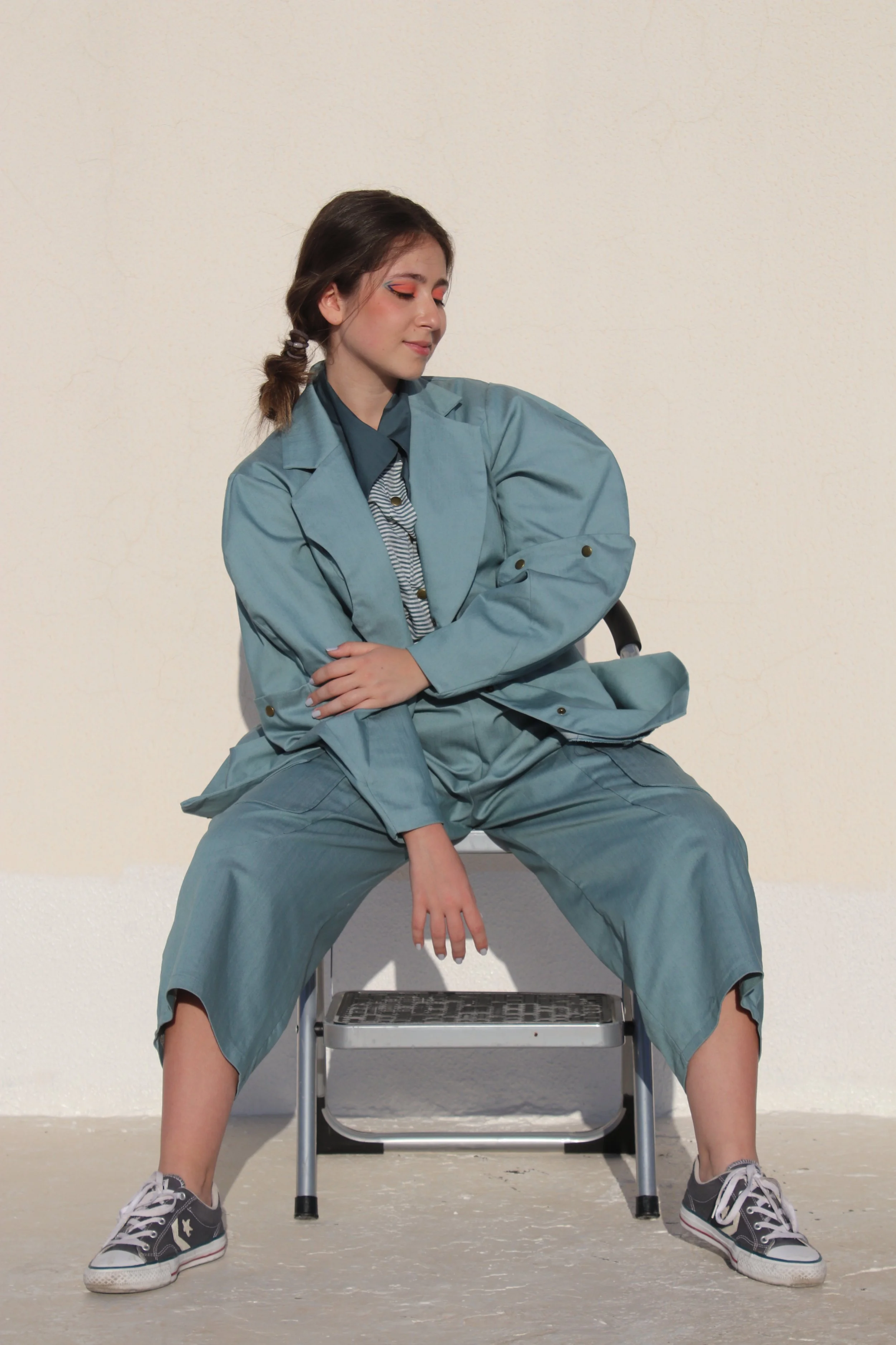 Young woman sitting on a chair with her legs apart, dressed in a teal suit and striped shirt, with makeup and hair styled casually, against a plain wall.
