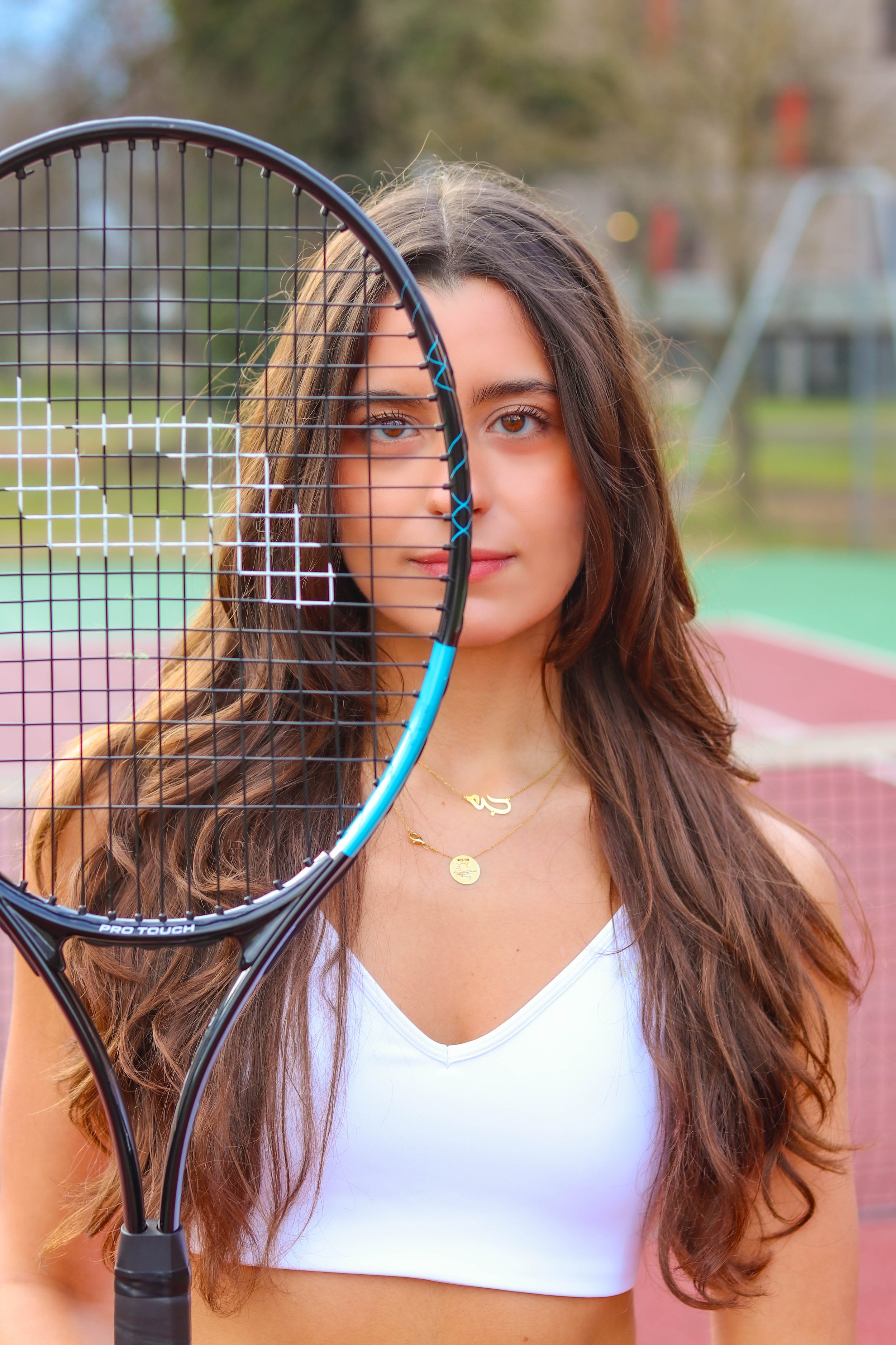 A young woman with long brown hair holding a tennis racket in front of her face, standing outdoors on a tennis court.