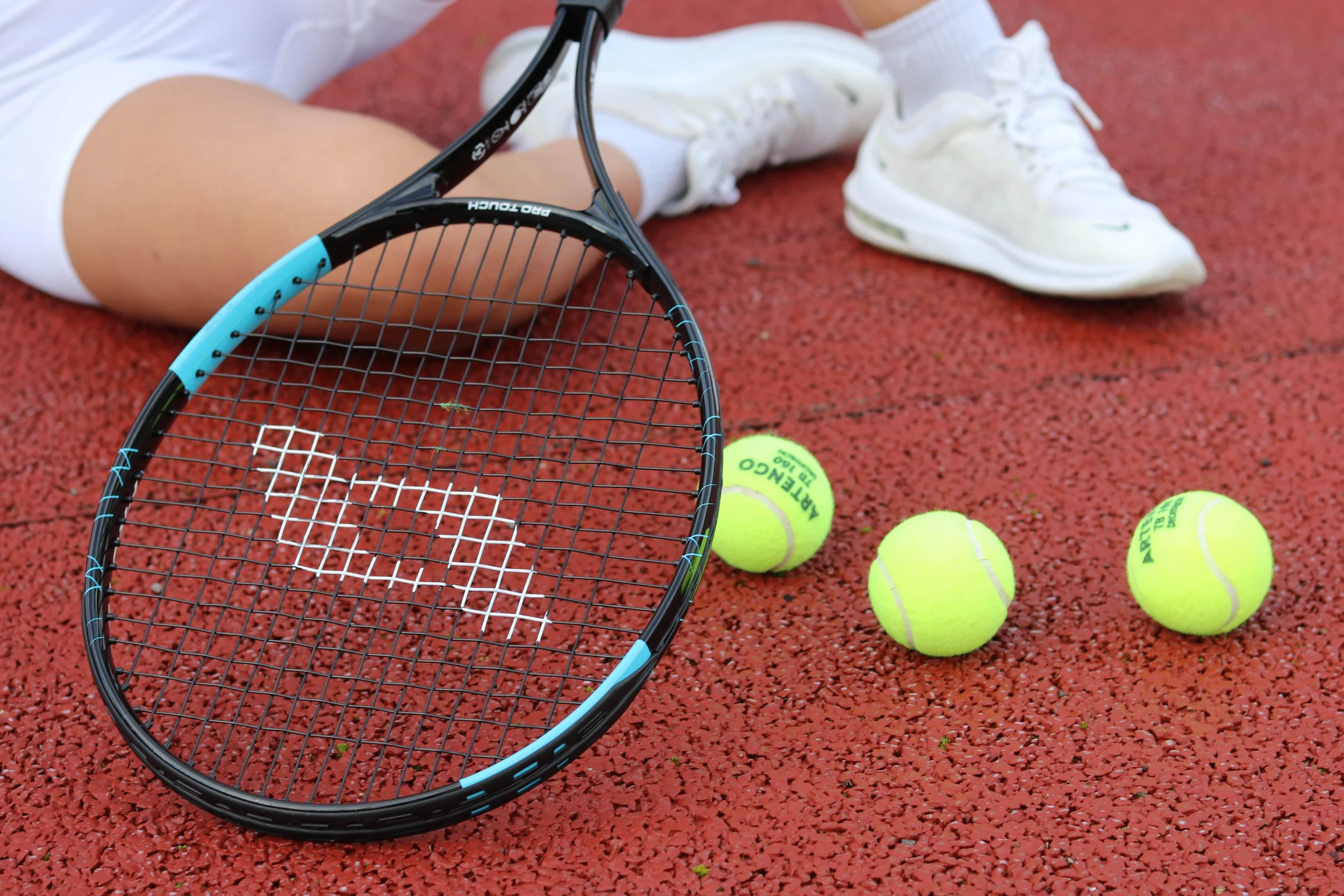 A tennis racket, tennis balls, and a person's legs on a reddish tennis court. The person is wearing white shoes and white socks, and is lying on the ground.