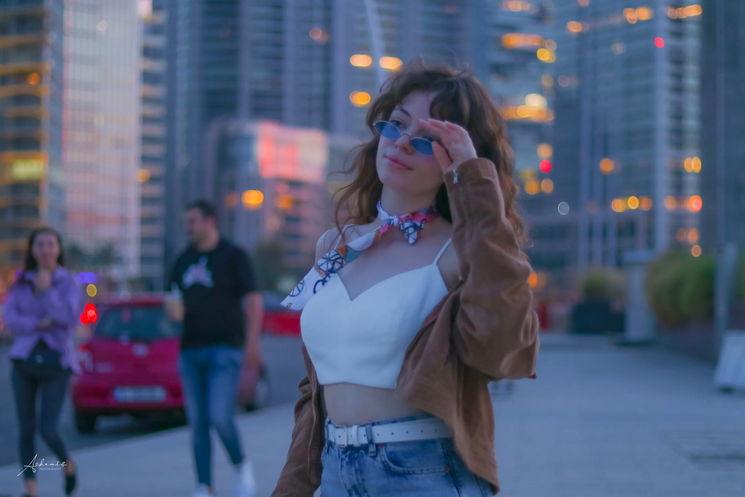 A young woman with curly hair, wearing blue sunglasses, a white crop top, a brown jacket, and jeans with a white belt, stands outdoors in front of tall modern buildings during sunset, adjusting her sunglasses.