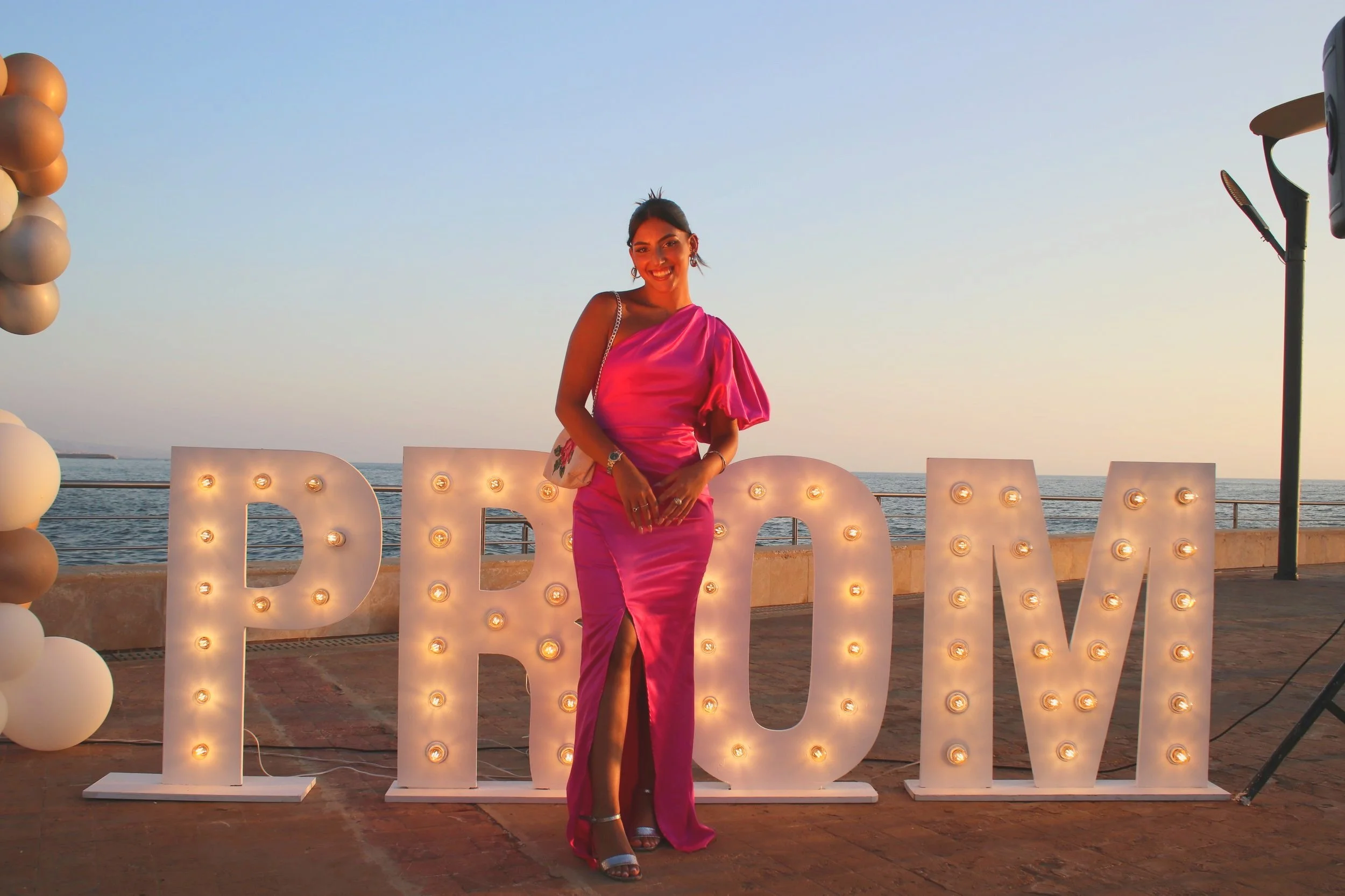 Woman in a pink satin gown standing next to illuminated letter signs spelling 'PROM' at a seaside location during sunset.