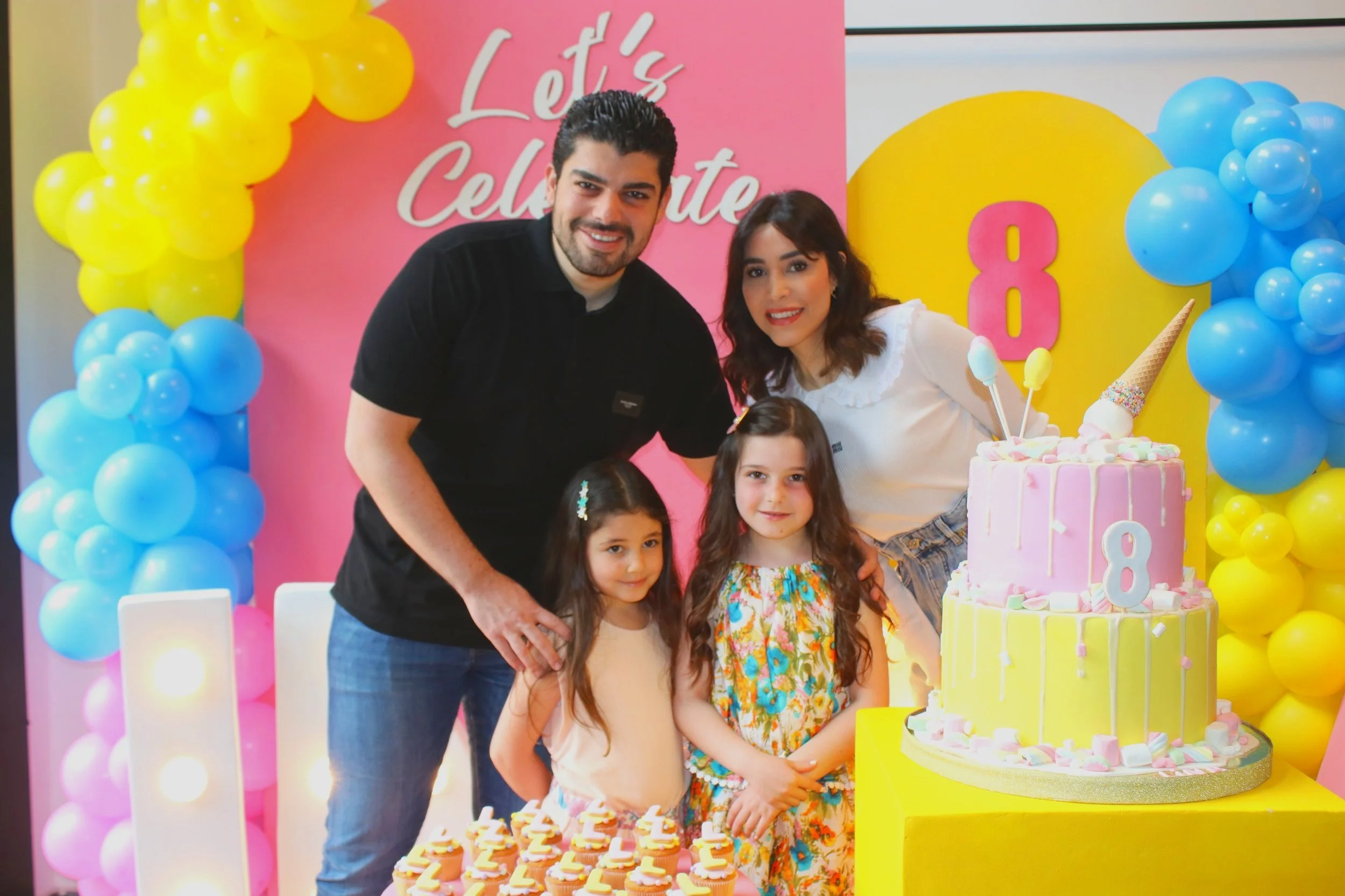 A family of four celebrating an 8th birthday with a pink and yellow themed party, including a pink cake, candy, and colorful balloons, with a pink backdrop that says 'Let's Celebrate.'