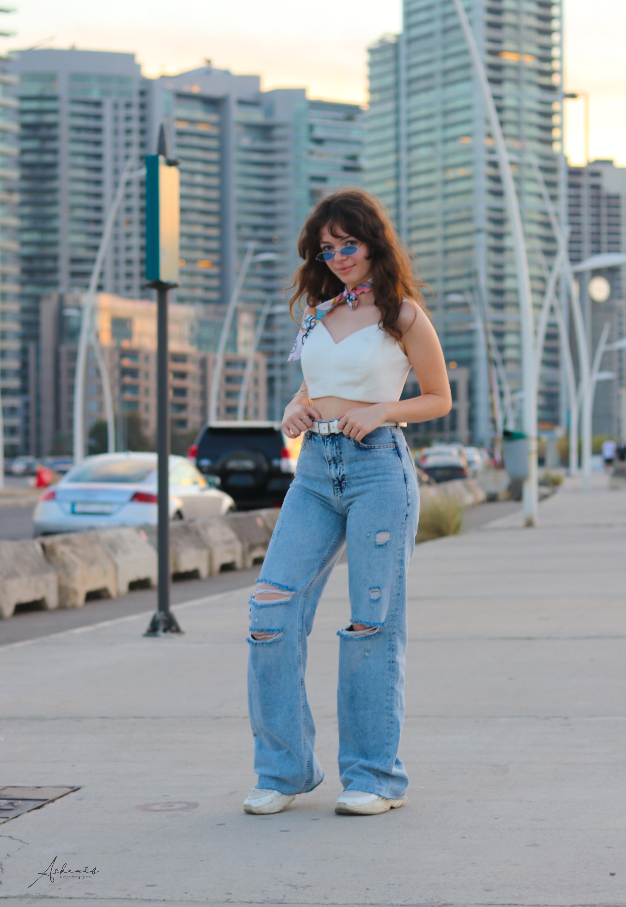 A young woman with curly brown hair and blue sunglasses standing on a city sidewalk at sunset, wearing a white crop top, baggy distressed jeans, and white sneakers.