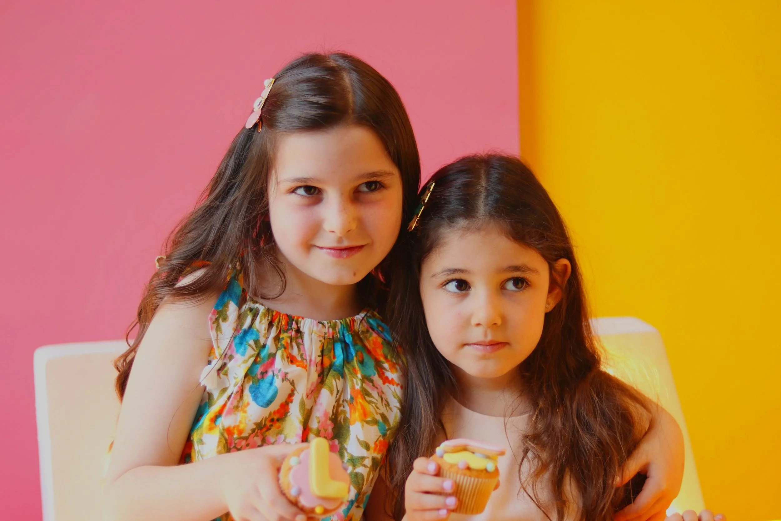 Two young girls holding cupcakes in front of a pink and yellow background.