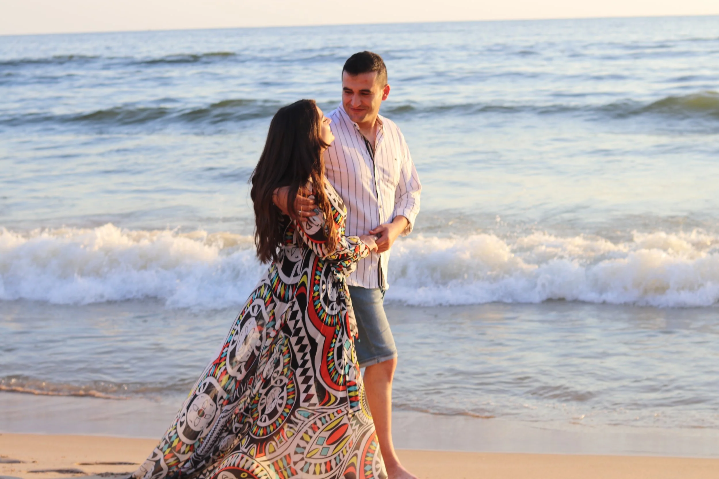 A young couple walking on the beach, holding hands, with waves in the background during sunset.