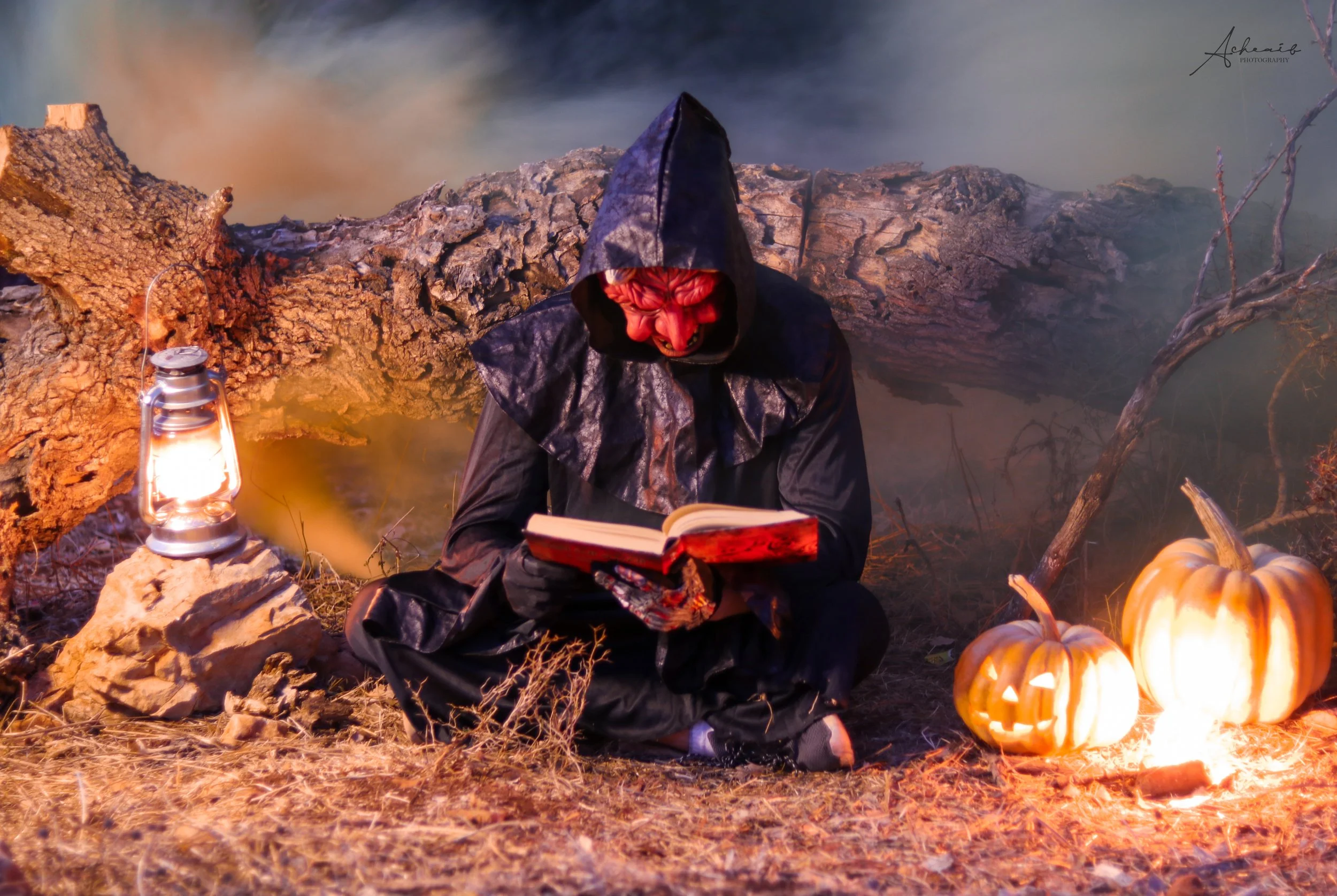 A person dressed as a witch in a black hooded cloak sitting on the ground by a tree, reading a book. There are two carved pumpkins with candles inside glowing, and a lantern on a rock nearby. The scene is set outdoors at dusk with autumn leaves and fog.