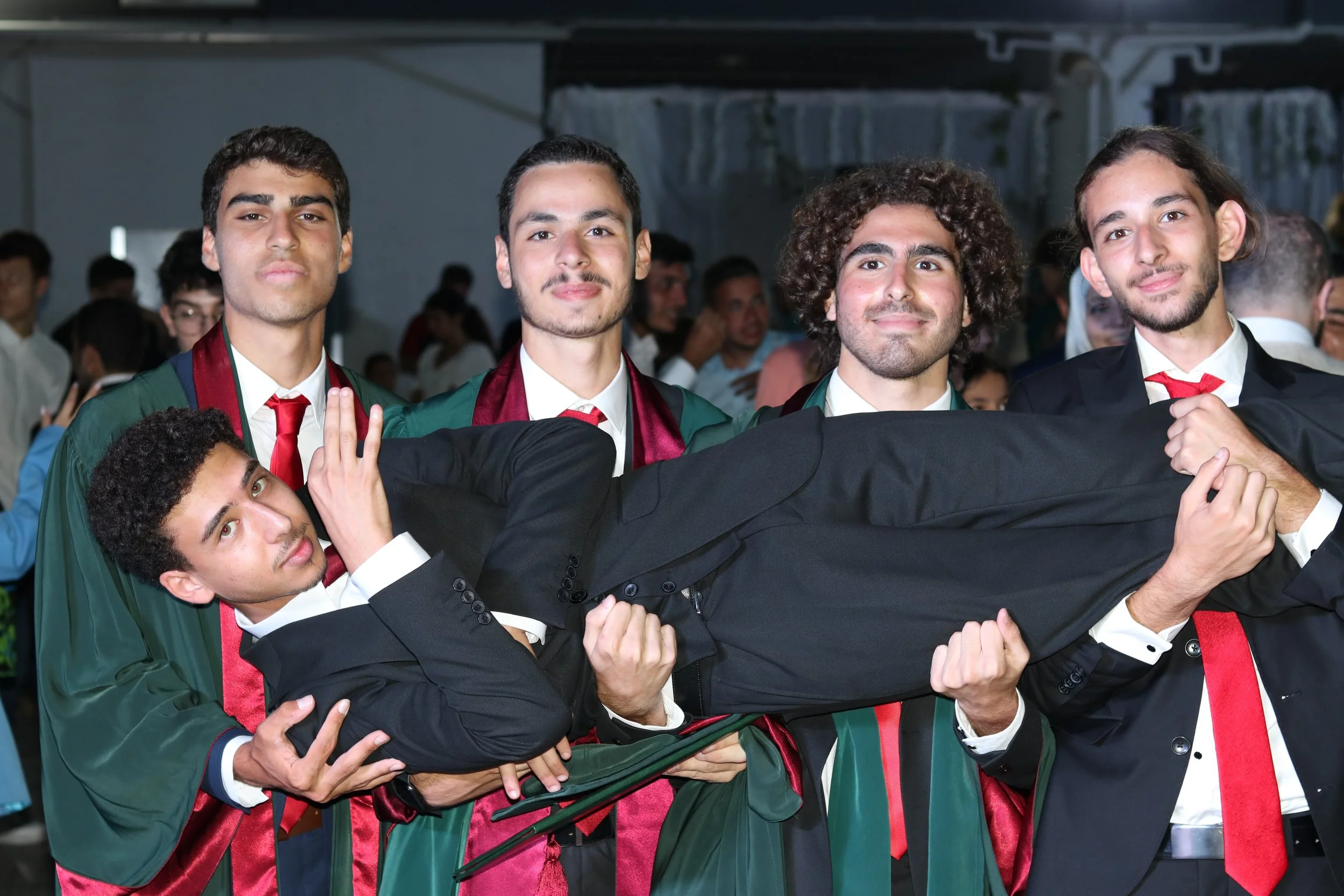 Group of five young men in graduation gowns, one lying horizontally, being carried by the others, celebrating graduation.