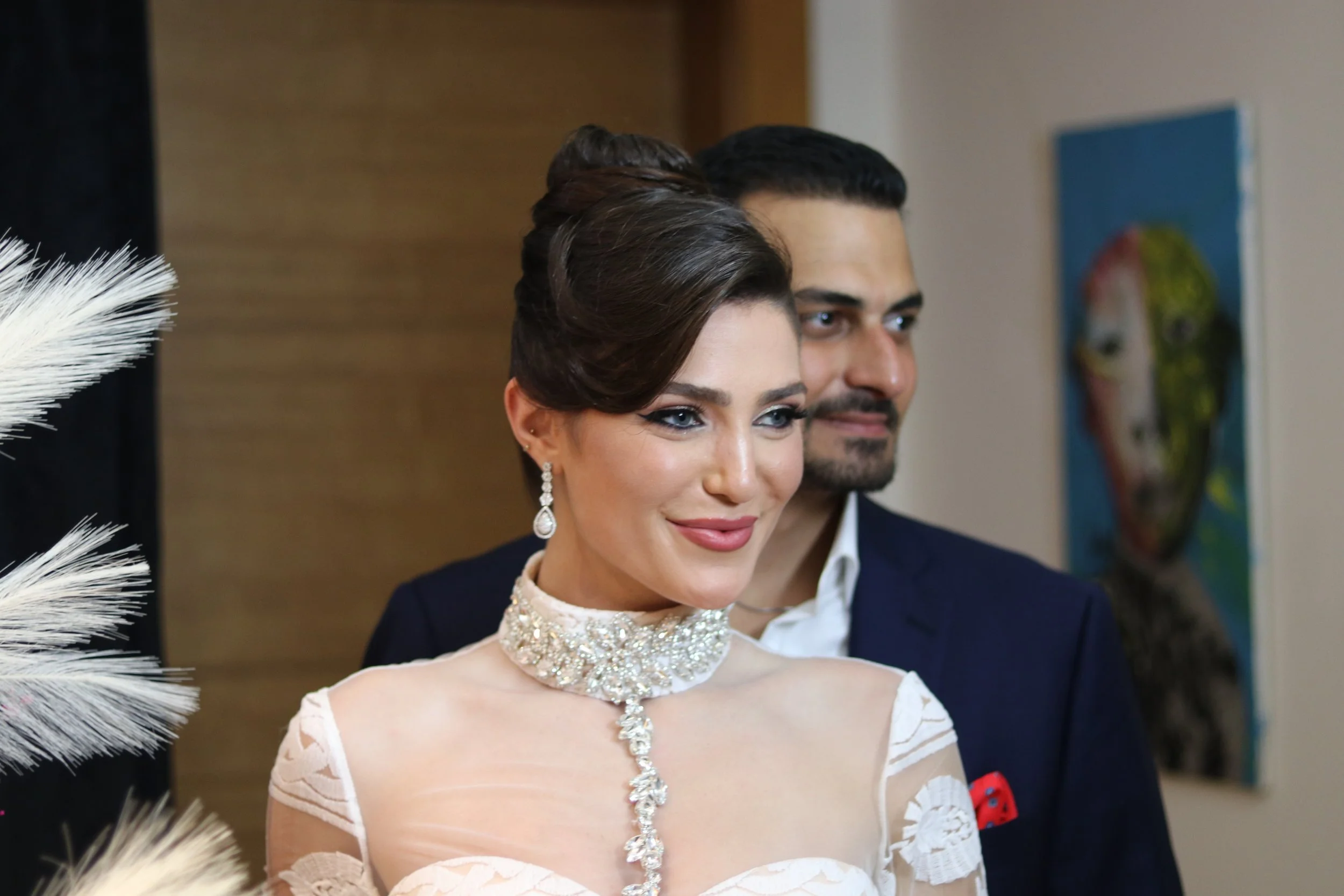 A woman with dark hair styled in an elegant updo and wearing glamorous jewelry, including a diamond necklace and earrings, stands in front of a man with dark hair and a beard. They are dressed formally, with the woman in a lace gown and the man in a navy suit, at what appears to be a social event or celebration.