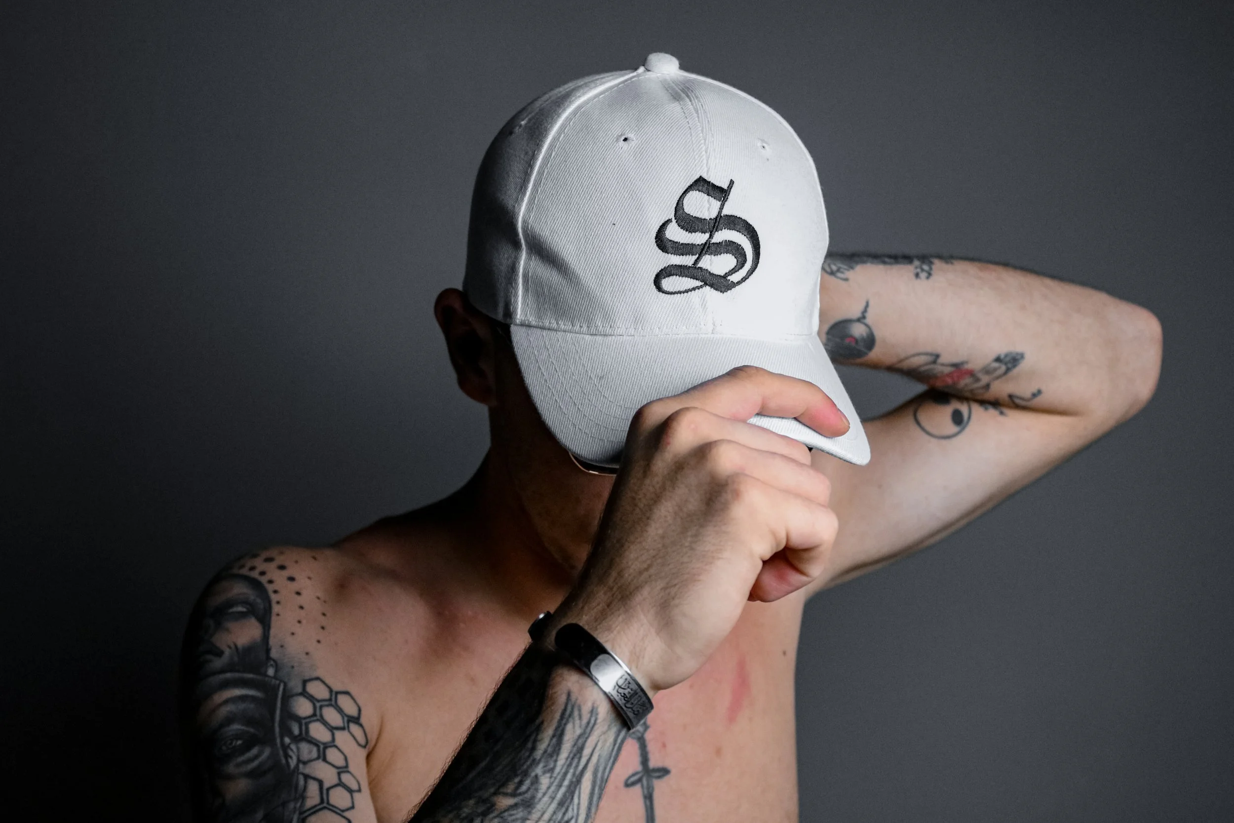 A shirtless person with tattoos wearing a white baseball cap with a black scripted logo, holding the brim of the cap with one hand and resting the other arm behind their head, against a gray background.