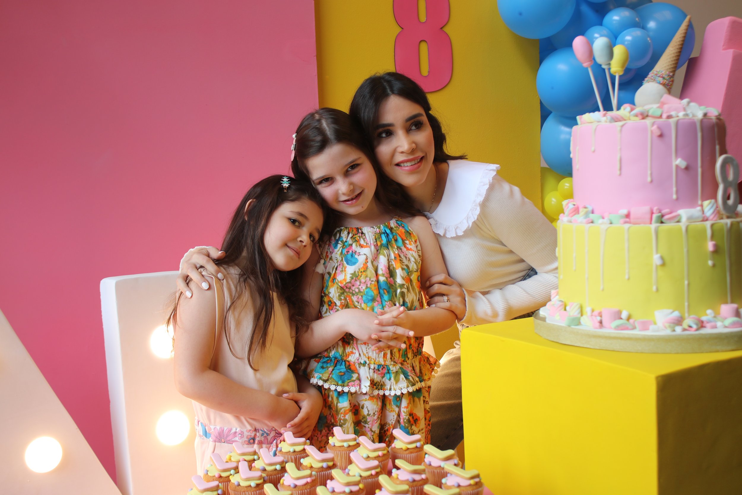 A woman and two young girls celebrating a birthday with a pink and yellow themed cake, cupcakes, and colorful decorations.