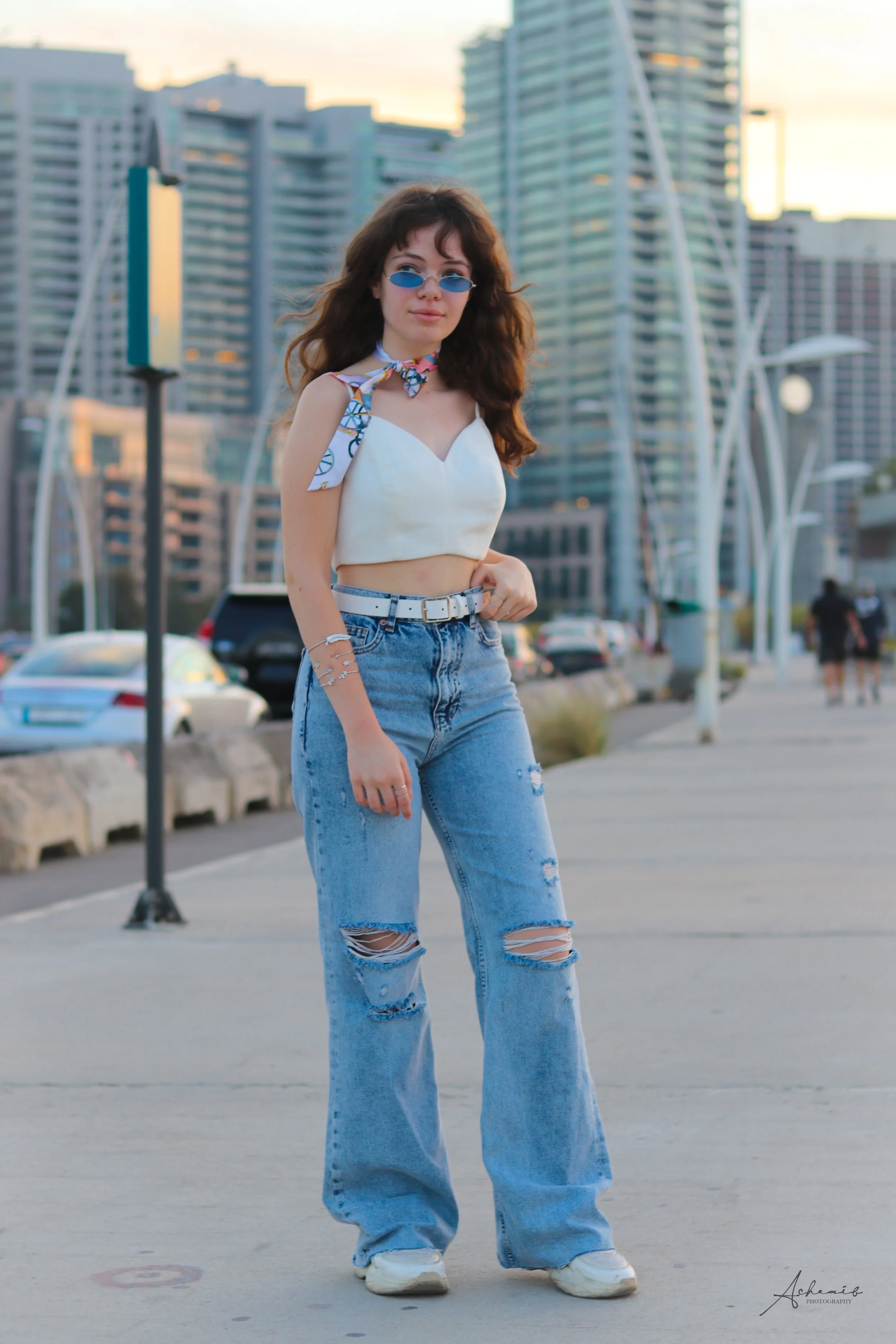 Young woman with curly brown hair wearing blue oval sunglasses, a white crop top, ripped baggy jeans, and a colorful neck scarf in an urban setting with modern buildings and parking lot.