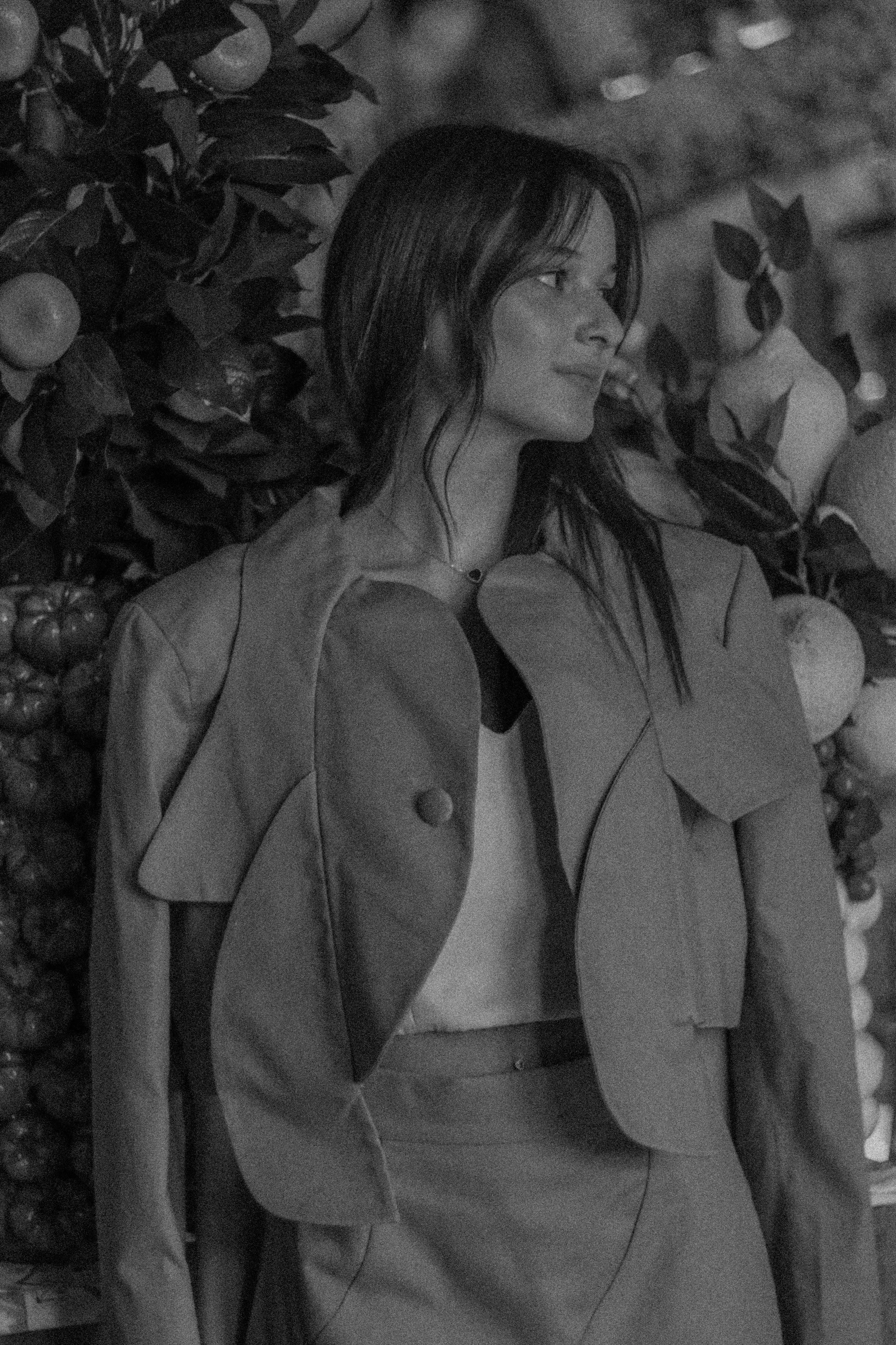 A woman with long dark hair is standing sideways among fruit trees, wearing a jacket and a skirt, in a black and white photo.