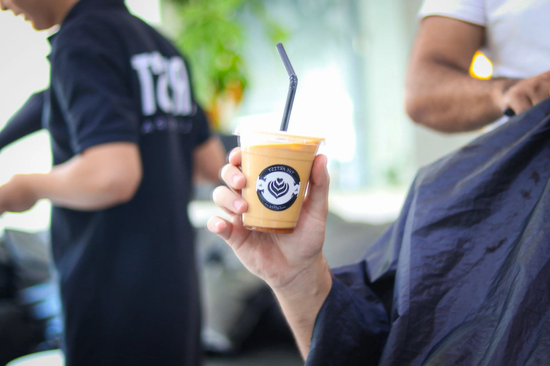 A person holding a plastic cup of iced coffee with a black straw, with a logo on the cup, in an indoor setting with another person in the background.