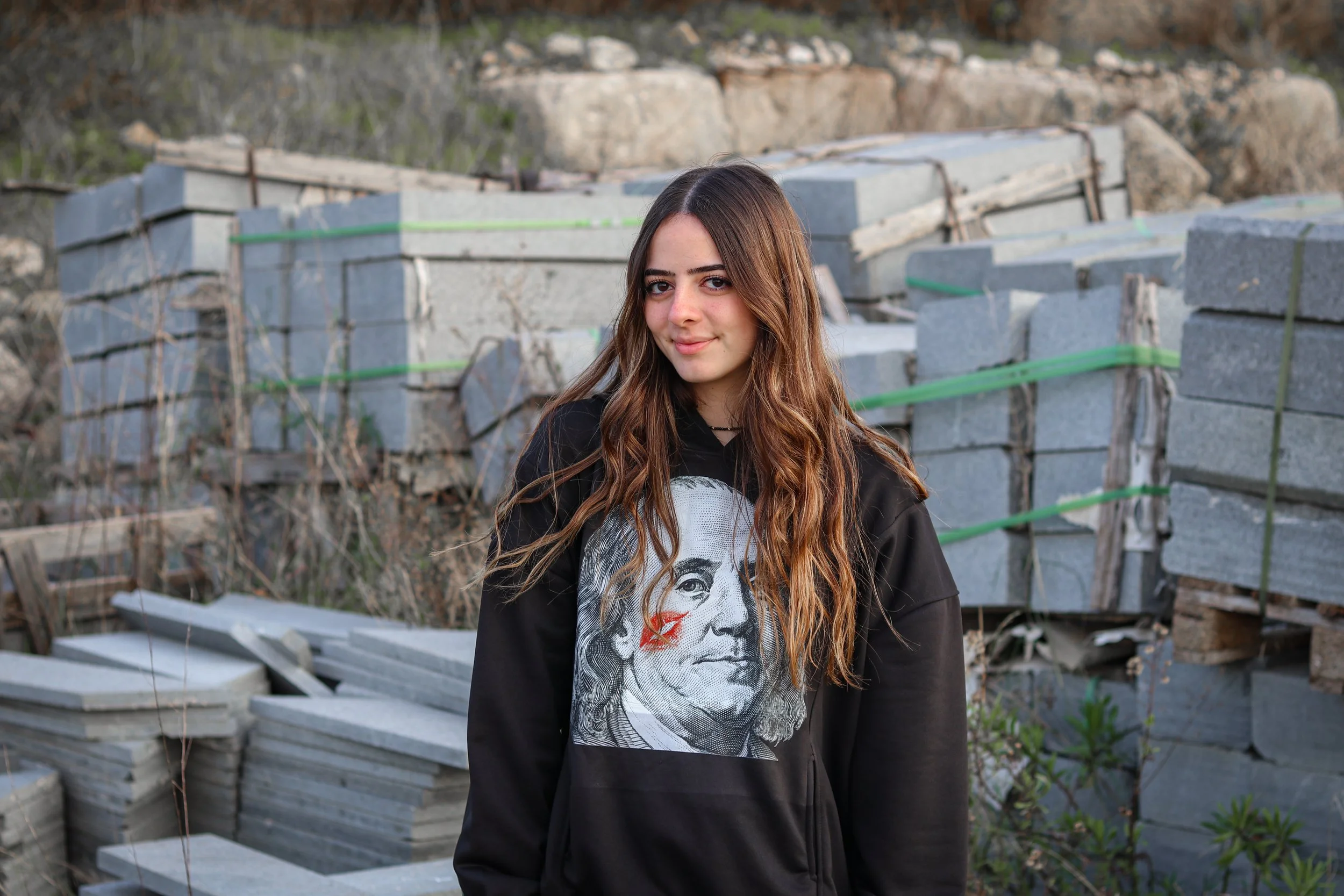 Young woman with long brown hair wearing a black hoodie with a portrait of Benjamin Franklin on it, standing in front of stacks of concrete blocks and rocks outdoors.