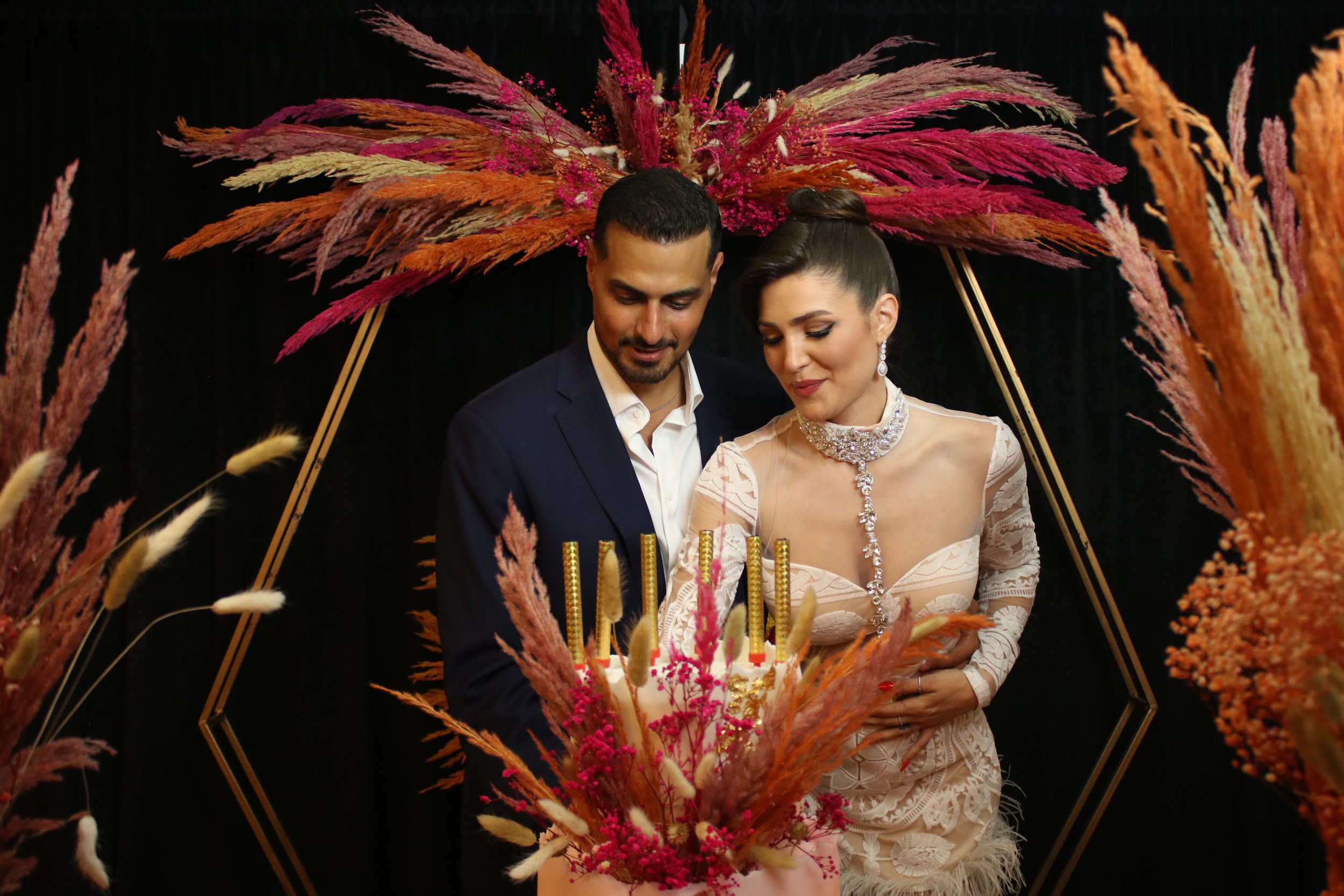 A newlywed couple at their wedding ceremony, standing behind a floral arrangement with pampas grass and pink accents, inside a decorative gold geometric frame, with a black backdrop.