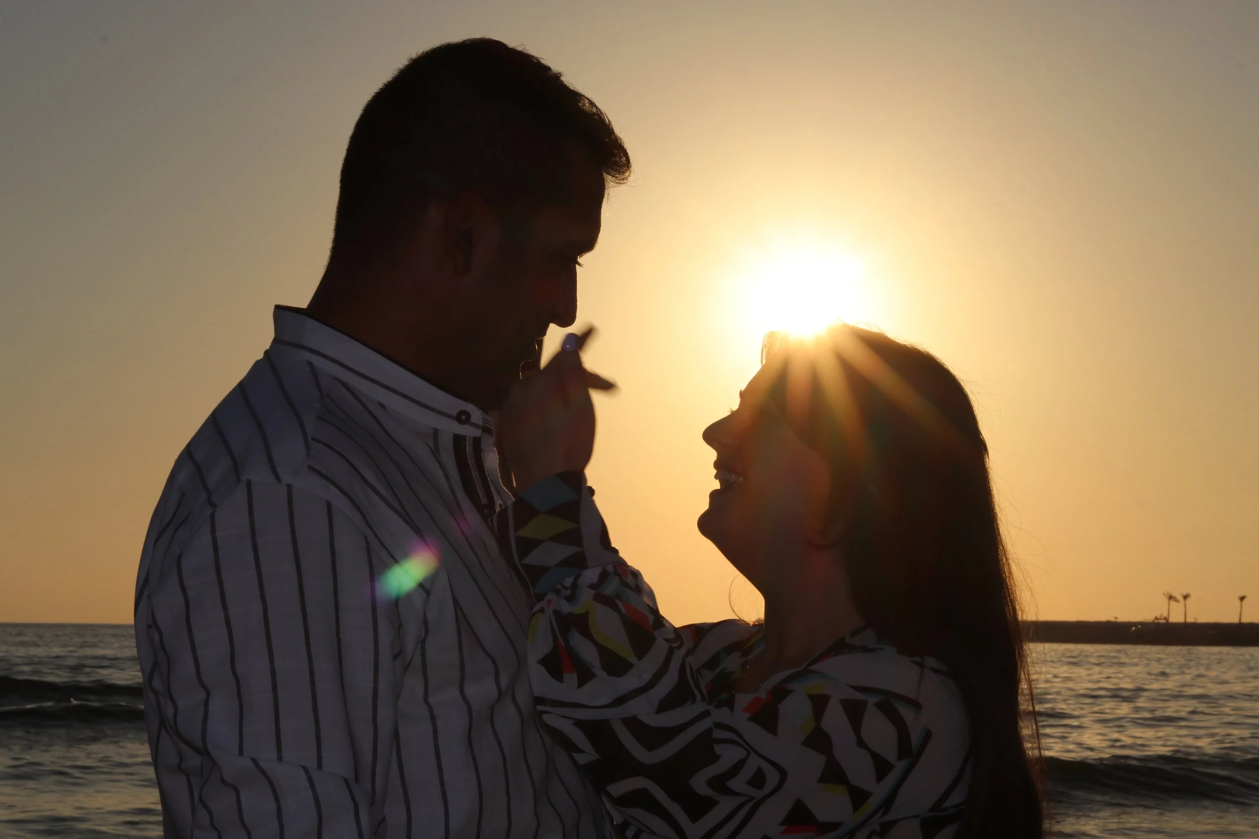 A silhouette of a couple facing each other on the beach during sunset, with the sun behind them creating a halo effect.