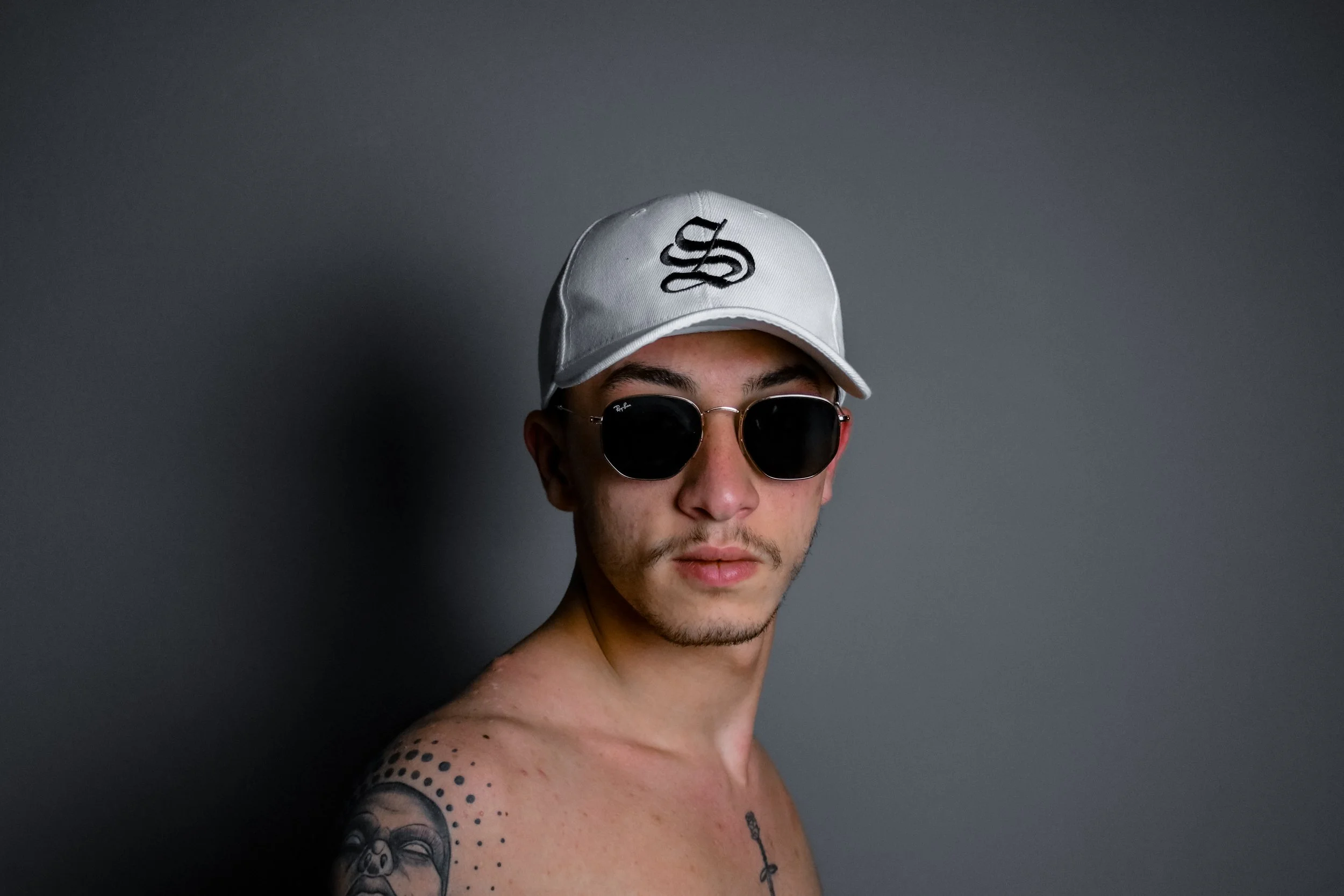 A shirtless man with tattoos wearing black sunglasses and a white baseball cap against a gray background.