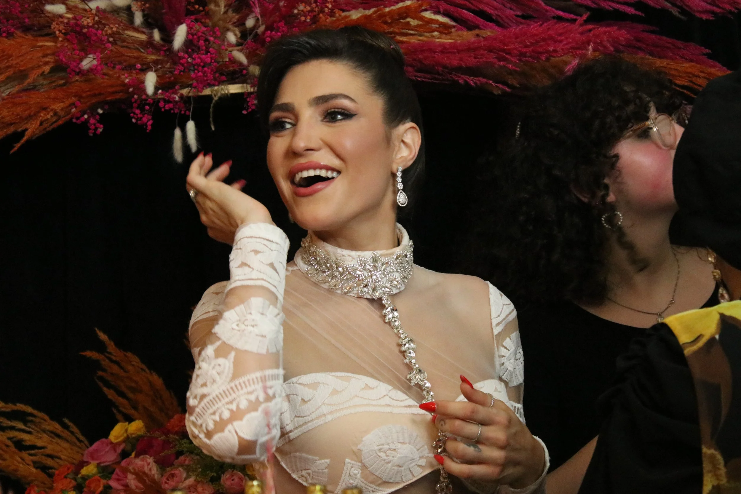 A woman dressed in an elegant, lace, white dress with intricate embroidery, wearing sparkling jewelry, including earrings, a necklace, and rings, smiling and touching her face at a celebration or event with colorful floral decorations in the background.