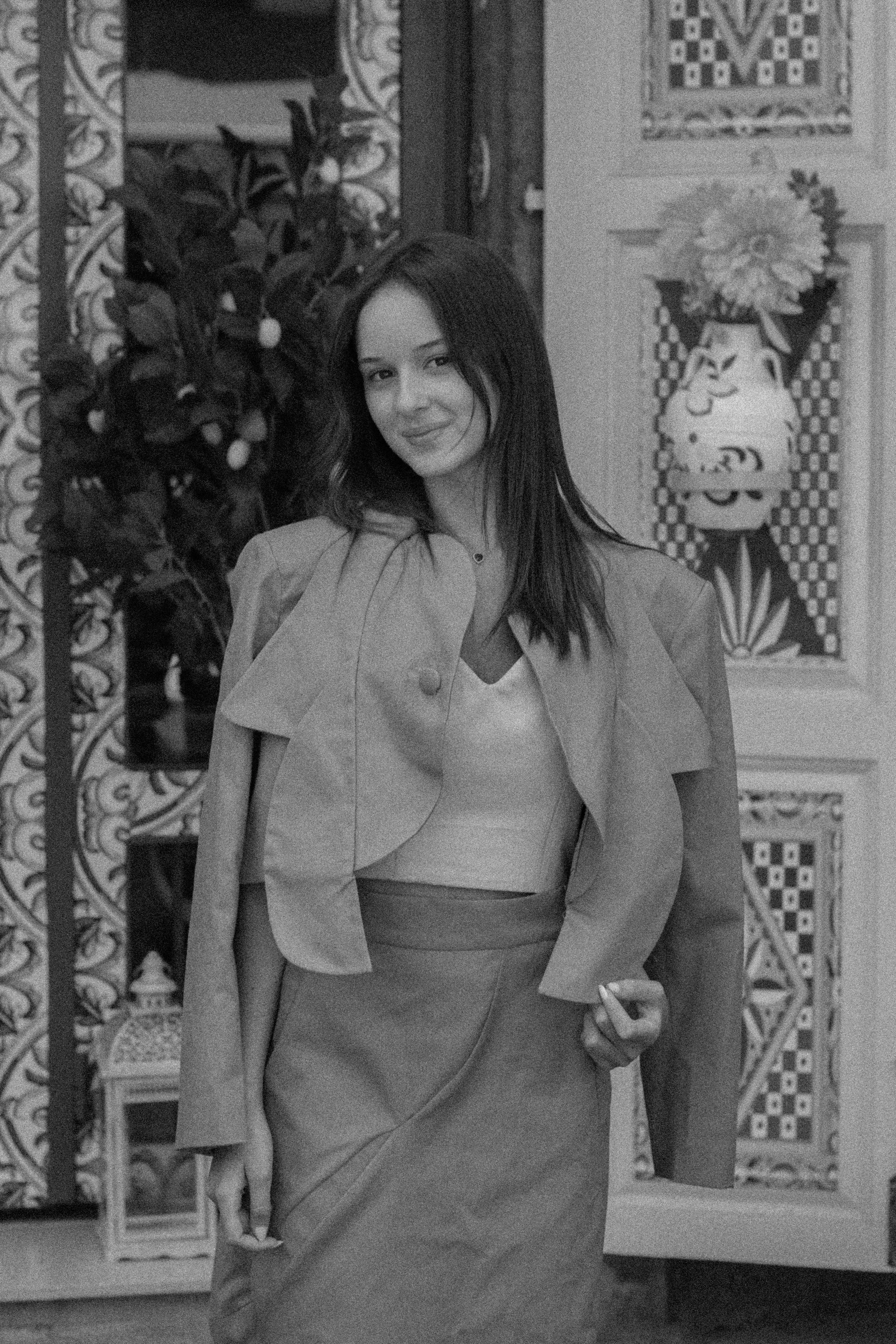 Young woman with dark hair smiling, standing indoors in front of decorated wall with floral patterns and art, wearing a blazer, blouse, and skirt.
