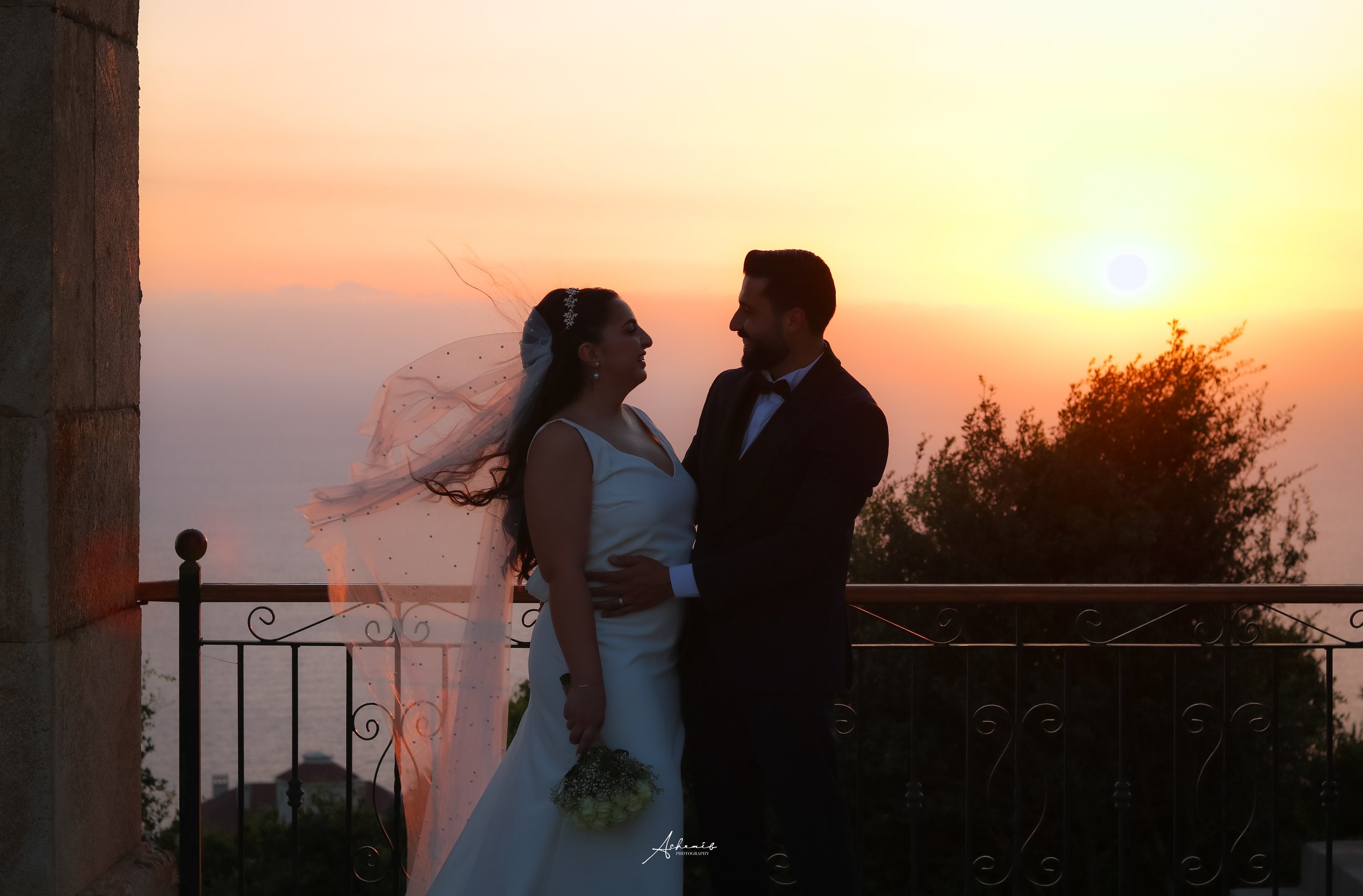 A bride and groom stand close together against a sunset background on a balcony, with the bride holding a small bouquet and wearing a veil, as they look at each other.