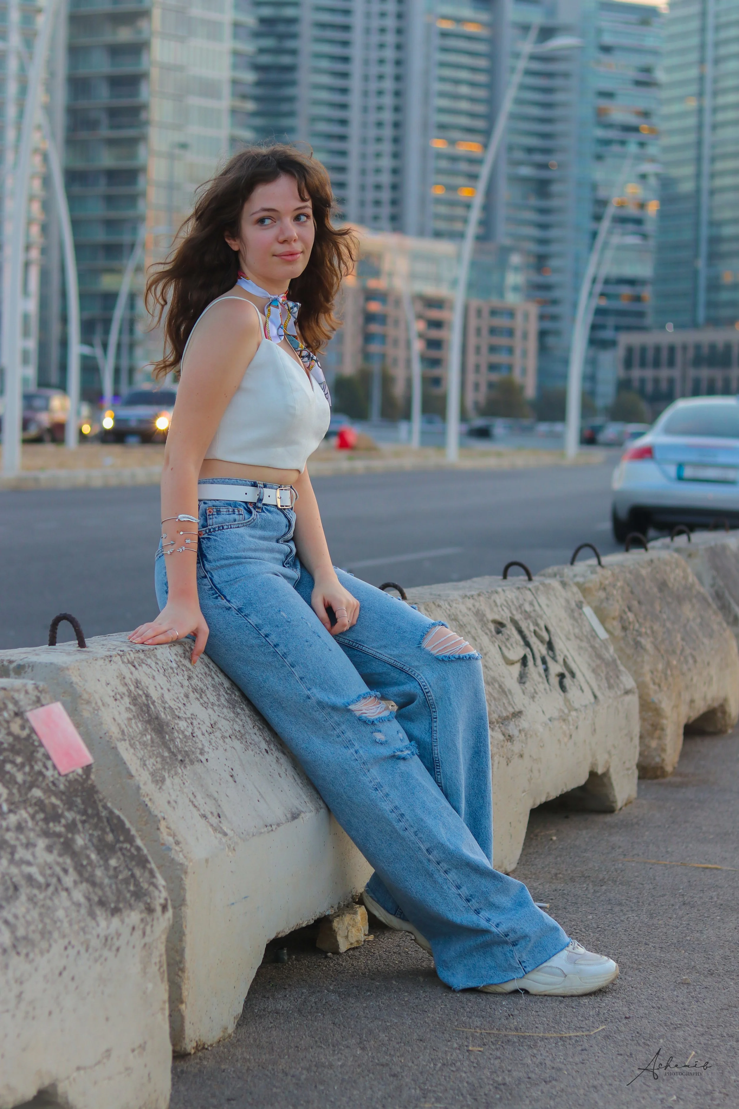 A young woman with long, wavy brown hair is sitting on a concrete barrier along a city street, with modern high-rise buildings in the background. She is wearing a white cropped top, ripped blue jeans, white sneakers, and a colorful scarf around her n