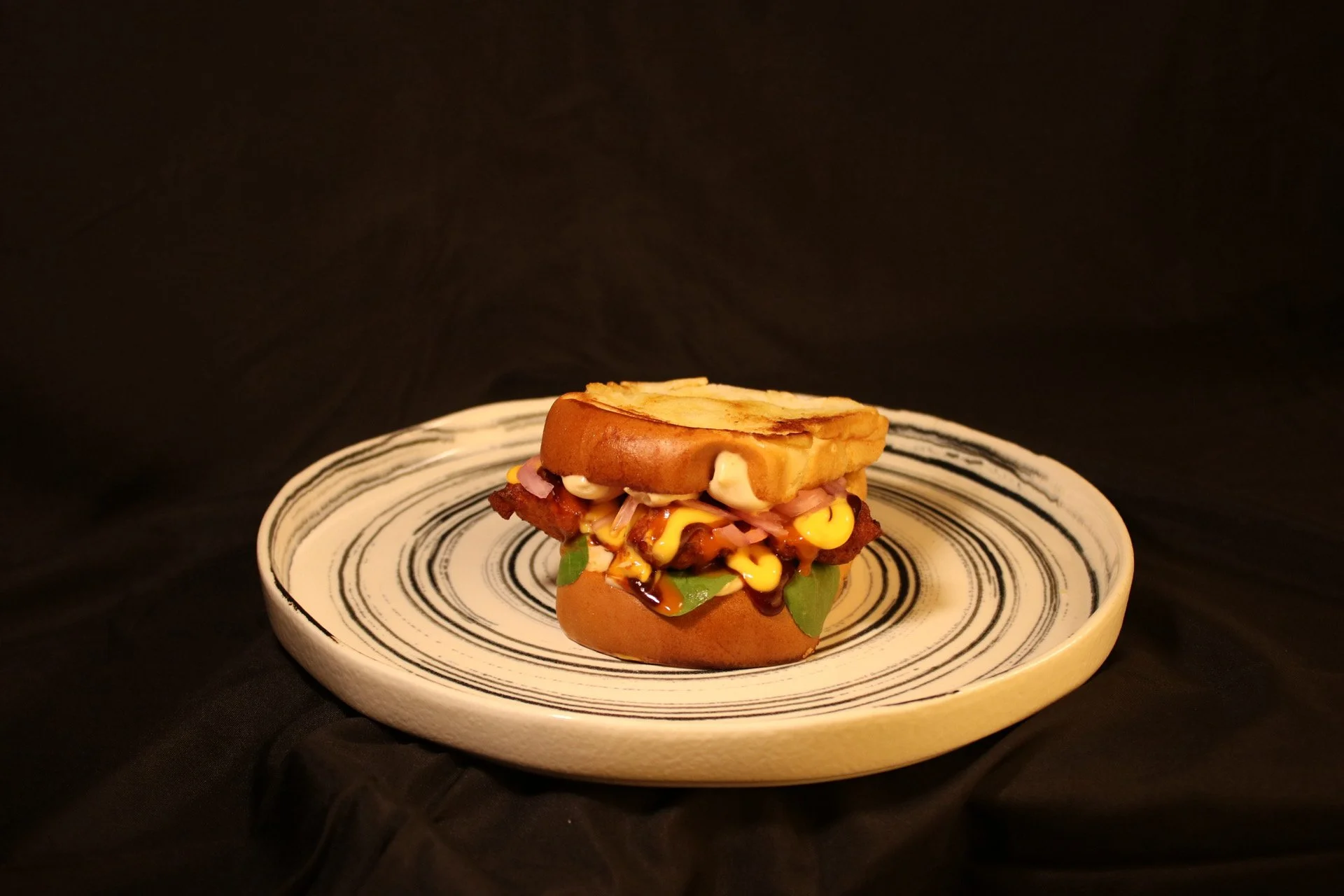 A burger with fried chicken fillet, cheese, pickles, onions, lettuce, sauce, and a toasted bun on a ceramic plate with black and white swirl design, against a black background.