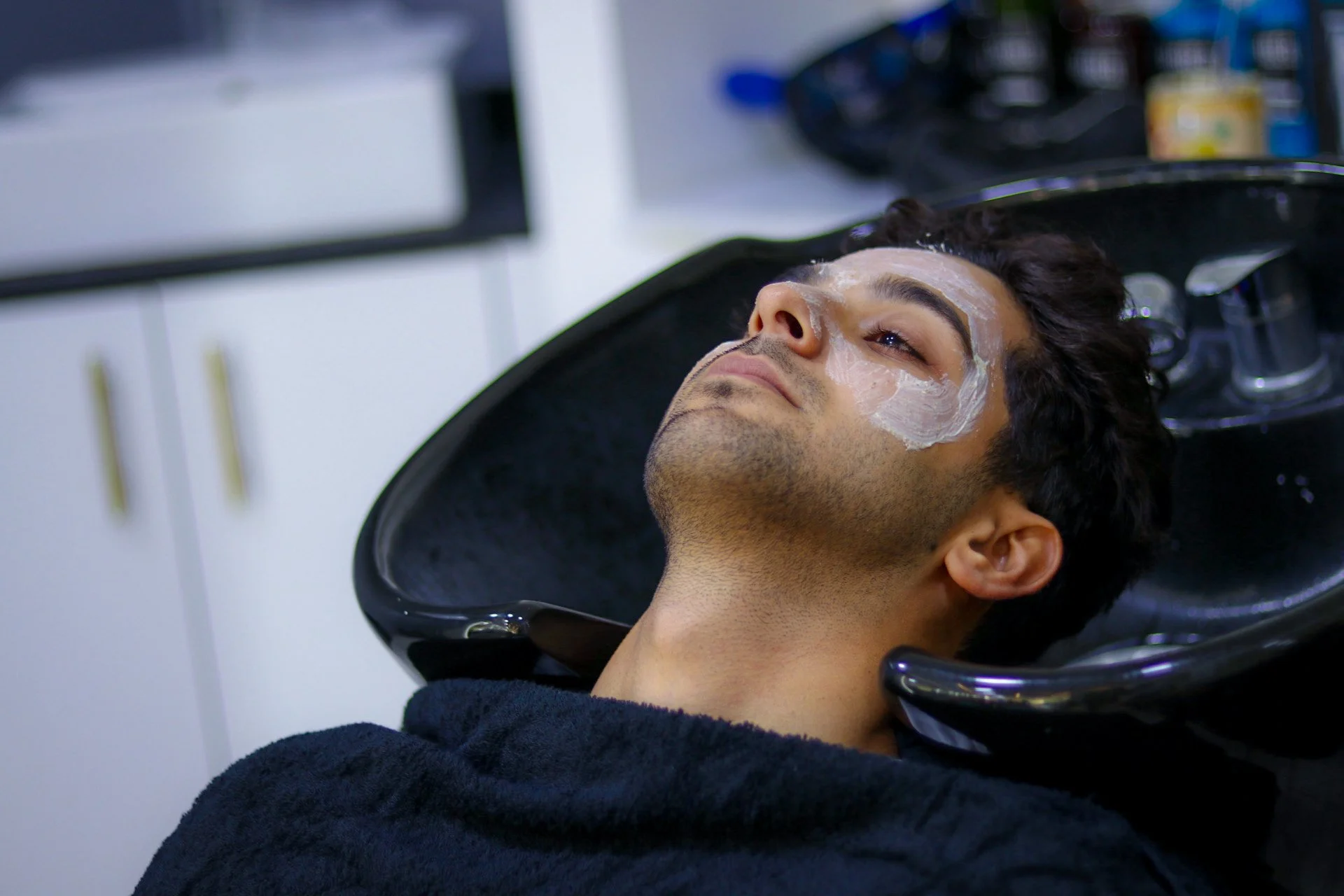 Man lying back with his head in a hair salon sink, getting his hair washed, with soap or shampoo on his face.