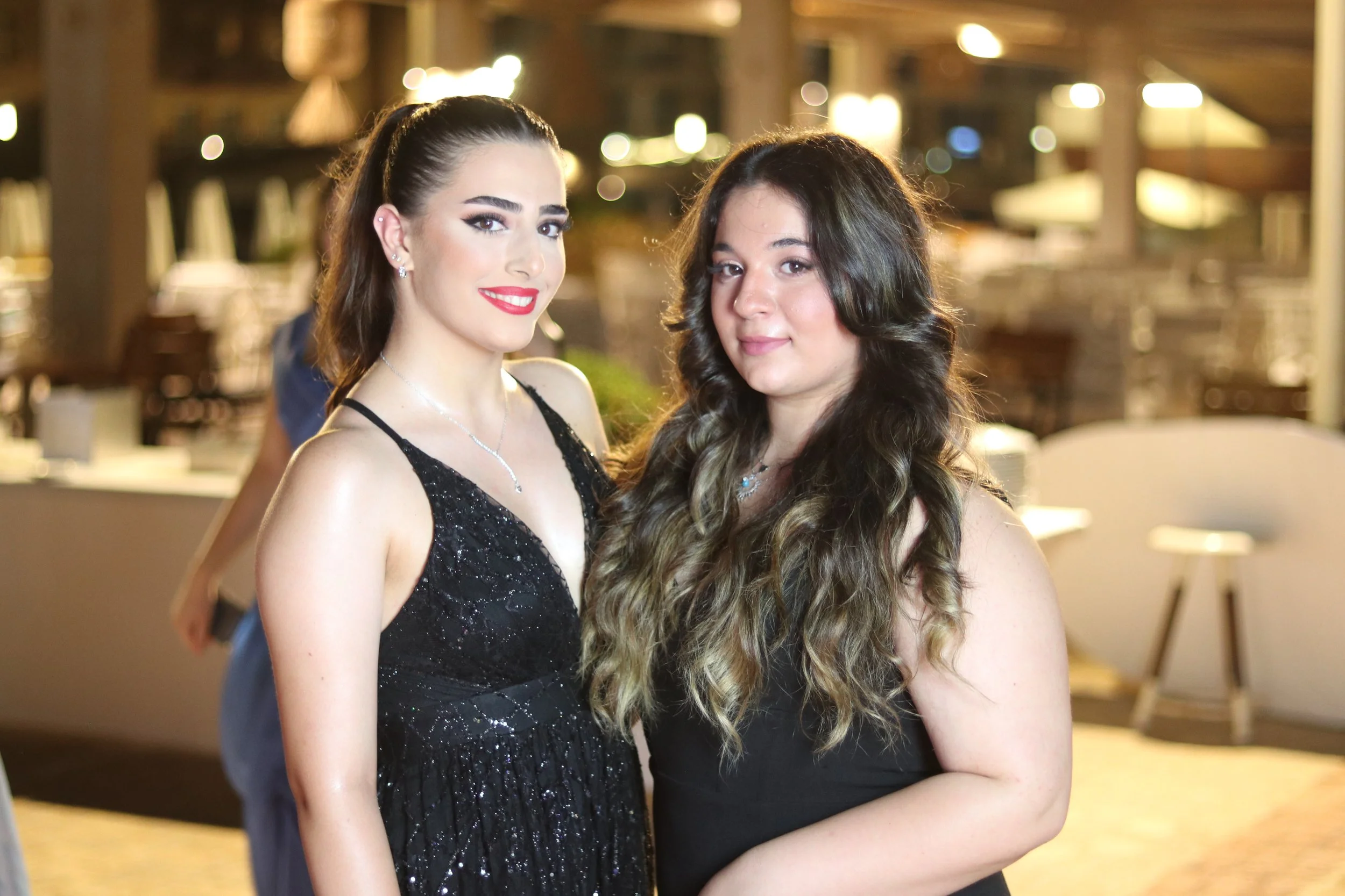 Two young women in black dresses standing close together at an indoor event, smiling at the camera, with blurred background lights.