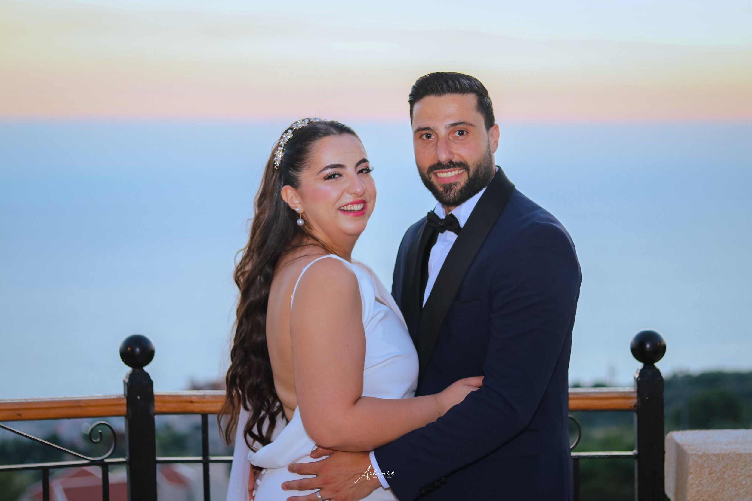 A newlywed couple in wedding attire embrace on a balcony overlooking the ocean at sunset.