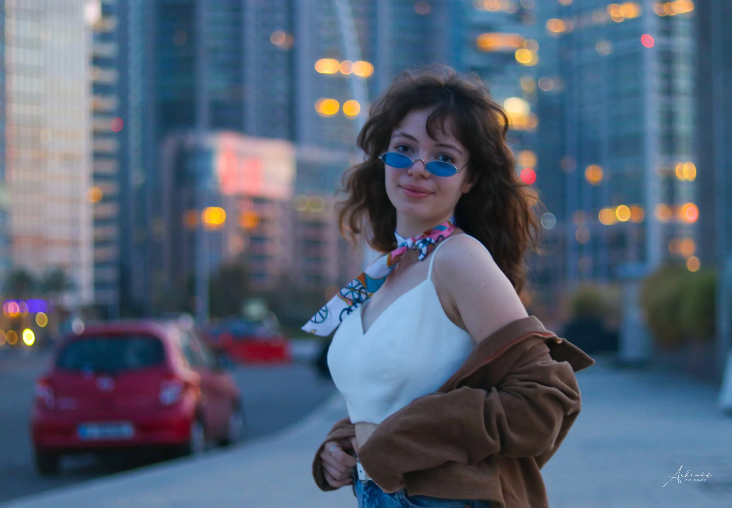 A woman with curly hair wearing blue sunglasses, a colorful scarf, a white sleeveless top, and a brown jacket draped over her shoulders standing outdoors in a city with blurred tall buildings and cars in the background during dusk.