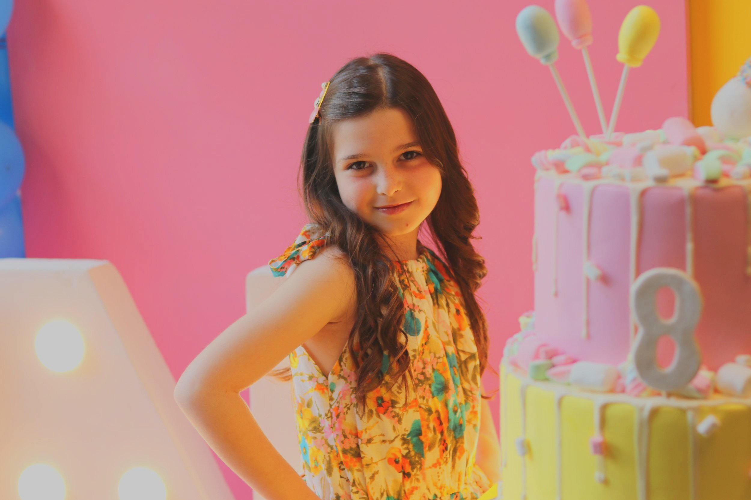 A young girl with long, wavy brown hair smiling at a birthday party. She is wearing a colorful floral dress and standing next to a pink and yellow birthday cake decorated with marshmallows and lollipops.