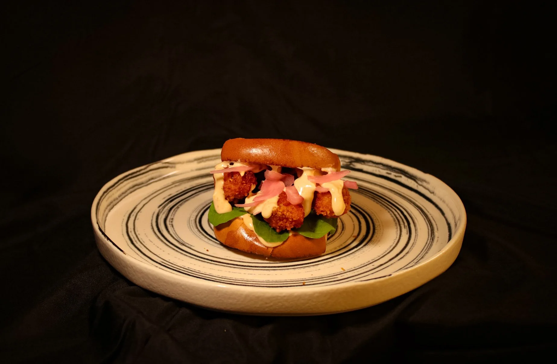 Fried chicken sandwich with lettuce, pink pickled onions, mayonnaise, on a toasted bun, served on a decorative plate against a black background.