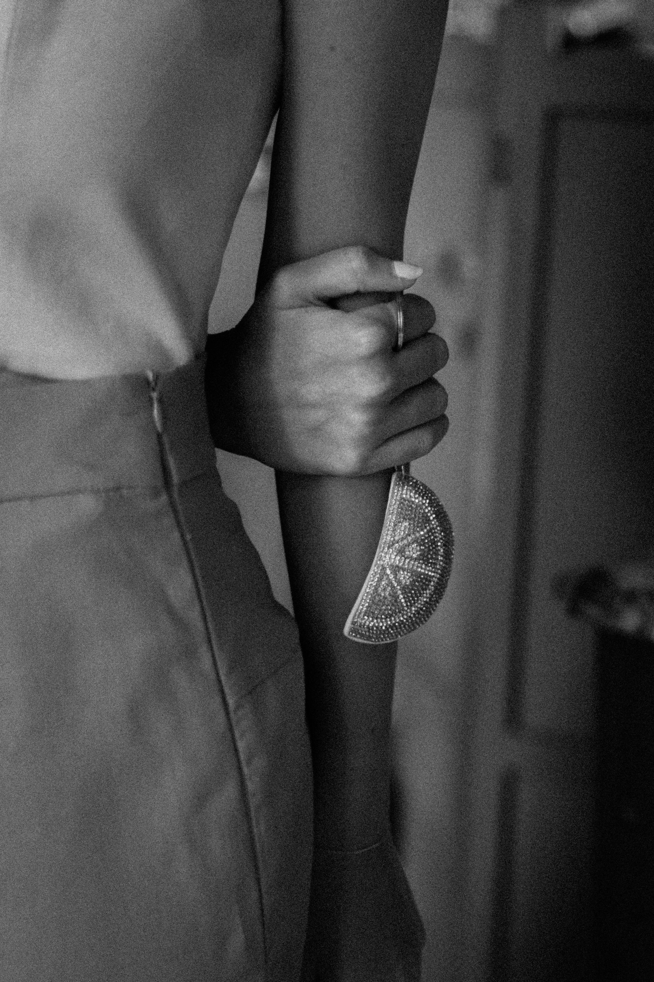 A black and white close-up photo of a woman’s hand gripping a pole, with a decorative piece hanging from her wrist and a small ring on her finger.