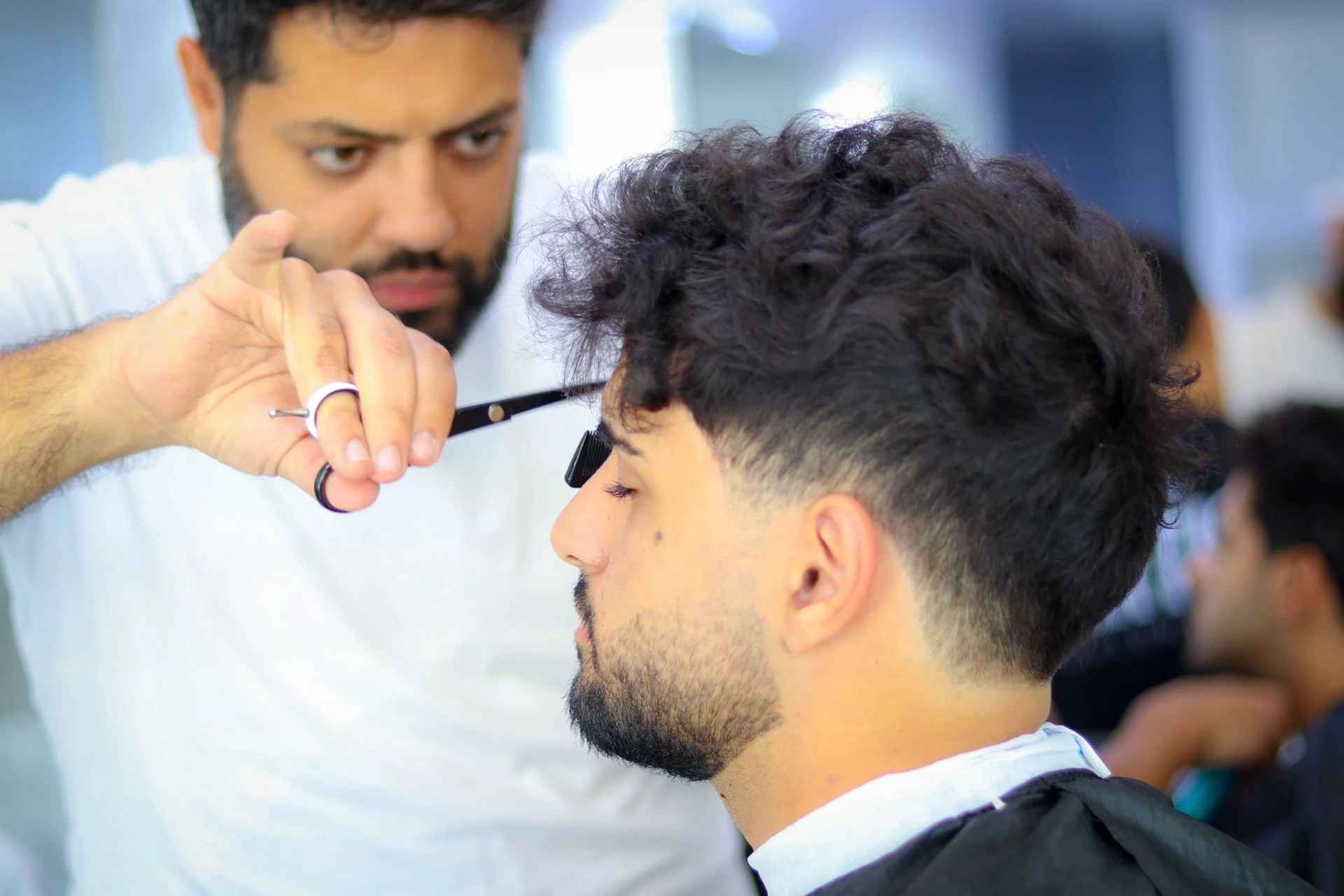 A hairstylist giving a young man a haircut in a salon, with other clients in the background.